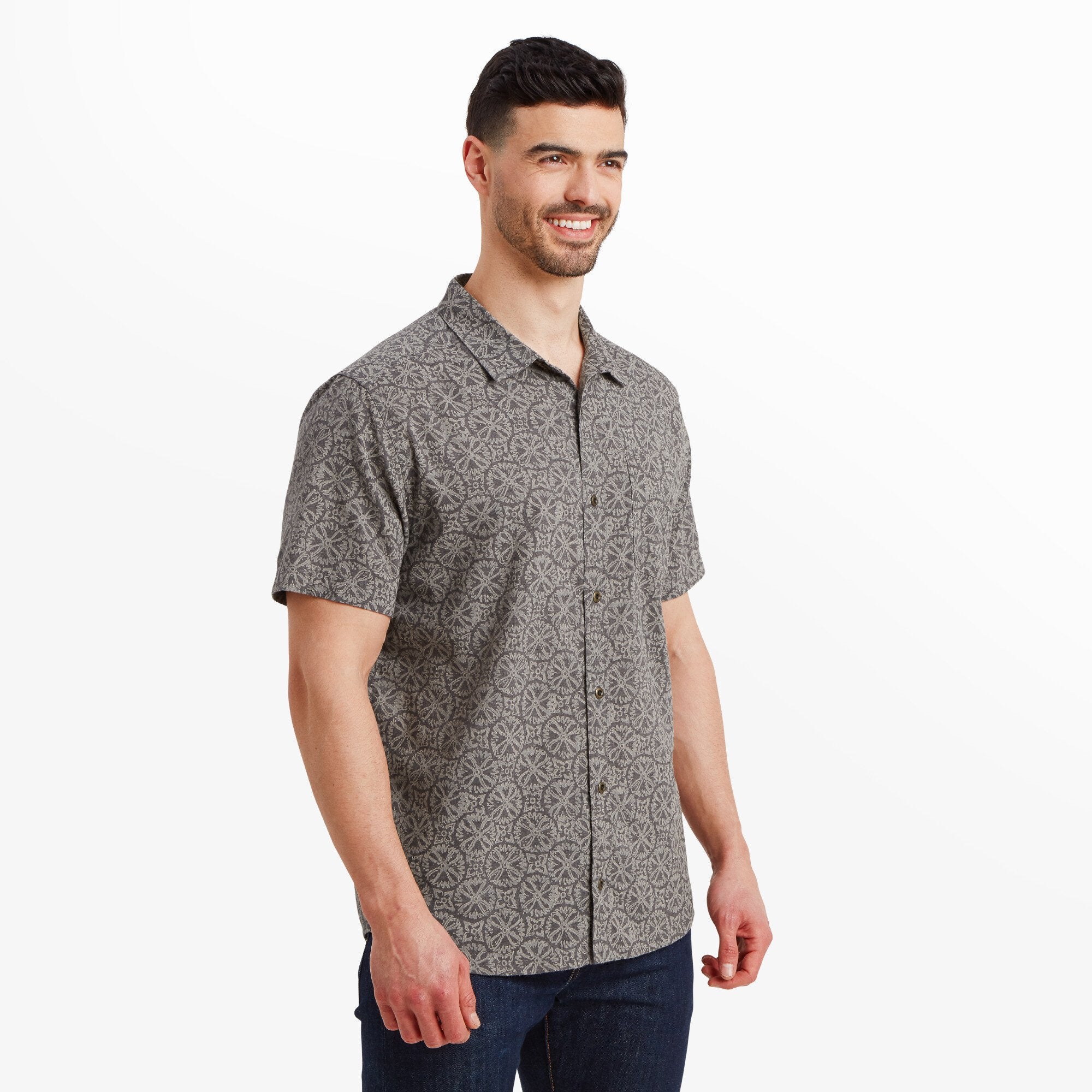 A man is modeling a Sherpa Adventure Gear Mancari Short Sleeve Shirt in Grey with a subtle grey geometric pattern. He is standing upright, smiling, and wearing dark jeans. The shirt has a pointed collar and a relaxed fit.