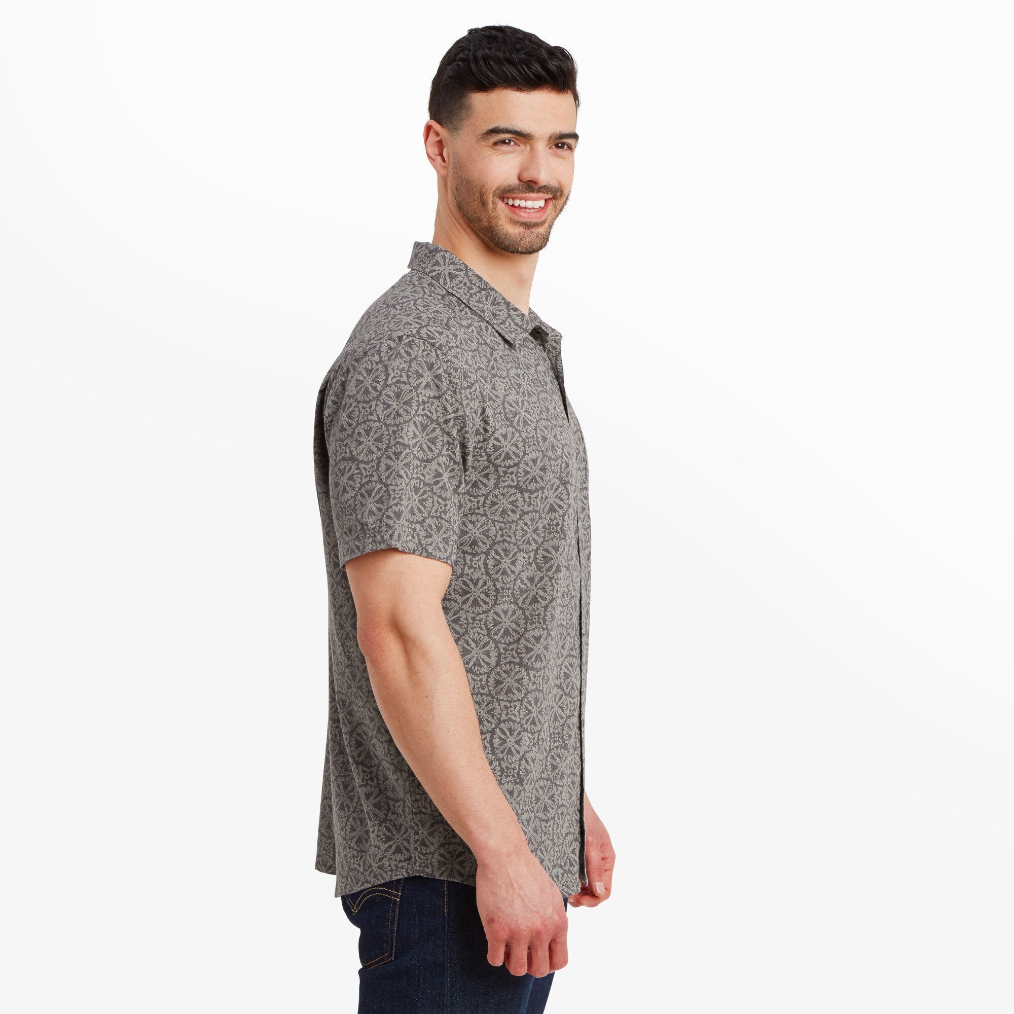 The man is facing to the side, showing the Sherpa Adventure Gear Mancari Short Sleeve Shirt in Grey's relaxed structure and the way it drapes on his torso. The geometric pattern is consistent throughout the fabric. The short sleeves fall slightly above his elbows.