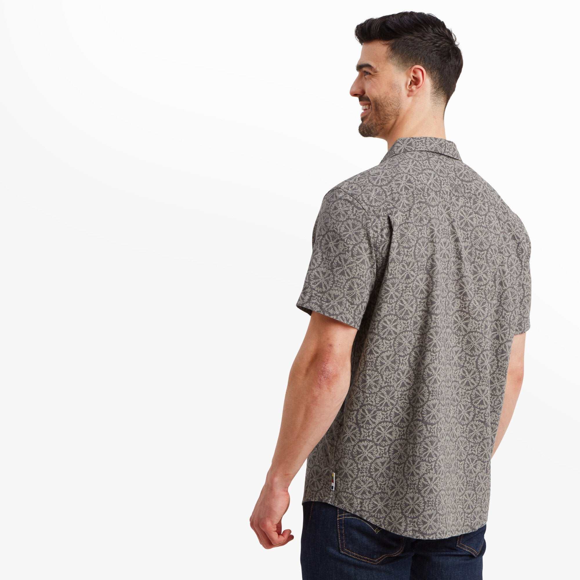 The man has turned his back to the camera, showcasing the Sherpa Adventure Gear Mancari Short Sleeve Shirt in Grey's back details. The geometric design covers the back evenly. The hem is slightly curved, offering a casual yet tailored look.