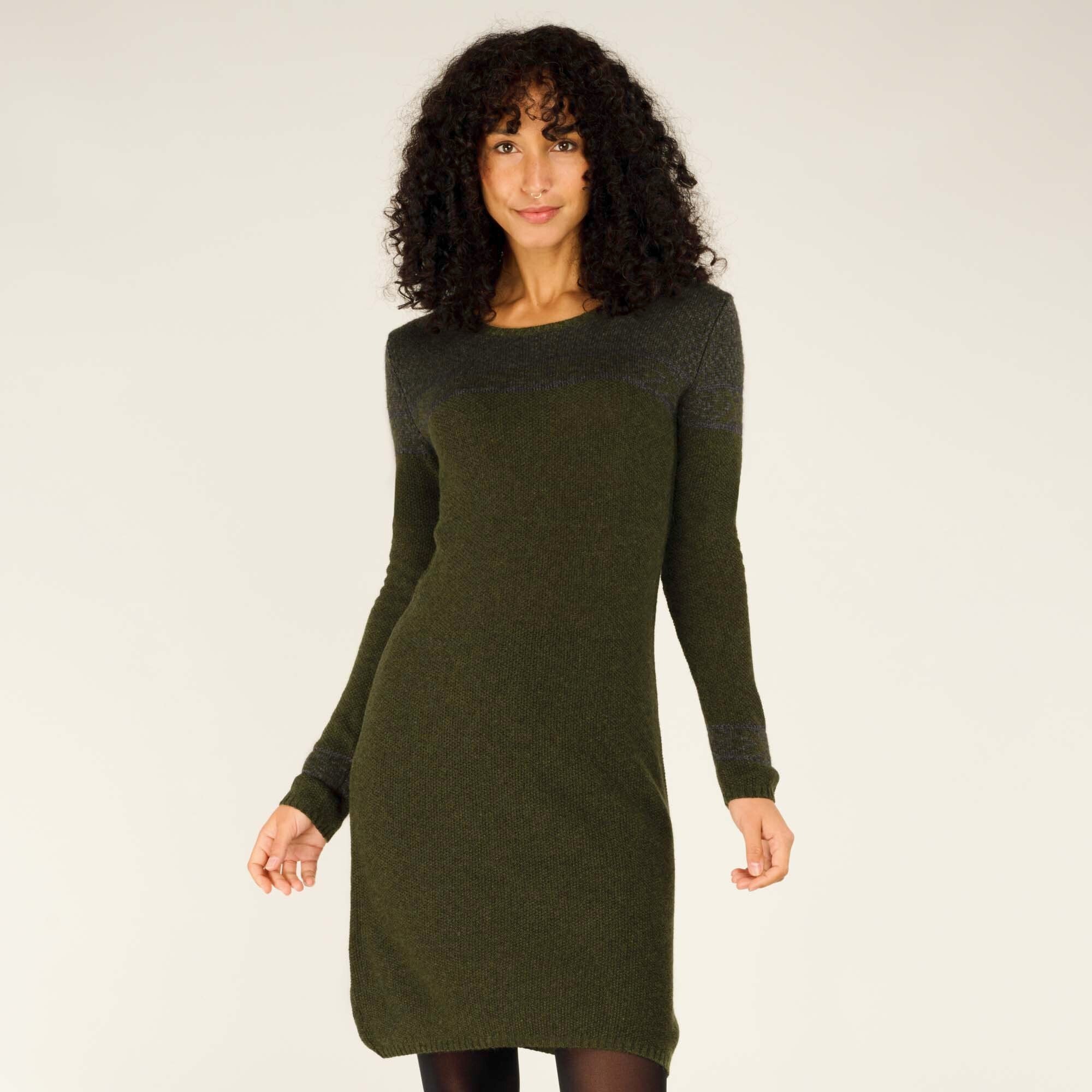 A woman with curly black hair, wearing a fitted Sherpa Adventure Gear Maya Texture Dress in Green. The dress has long sleeves with subtle horizontal knit details near the shoulders and cuffs. She stands with one hand at her side and the other slightly raised, smiling softly. The background is a neutral beige tone.