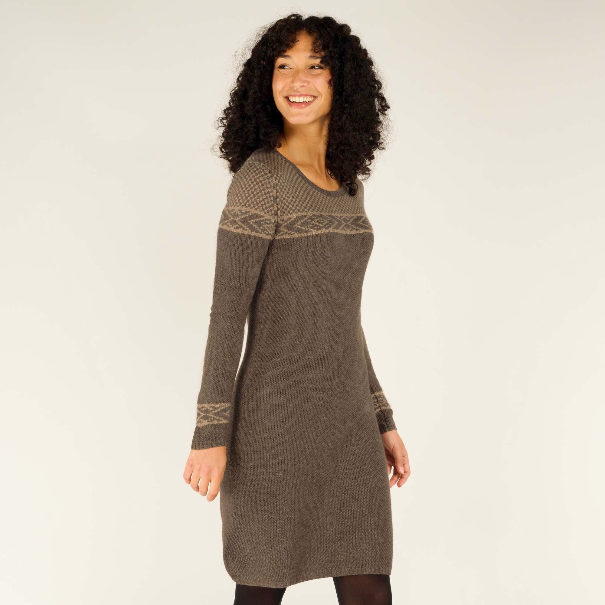 A smiling model wearing a Sherpa Adventure Gear Maya Texture Dress in Grey with intricate geometric patterns across the chest and cuffs. The dress has a fitted silhouette and ends just above the knees. The model is standing at a slight angle, looking over her shoulder with a cheerful expression, and the background is a neutral tone.