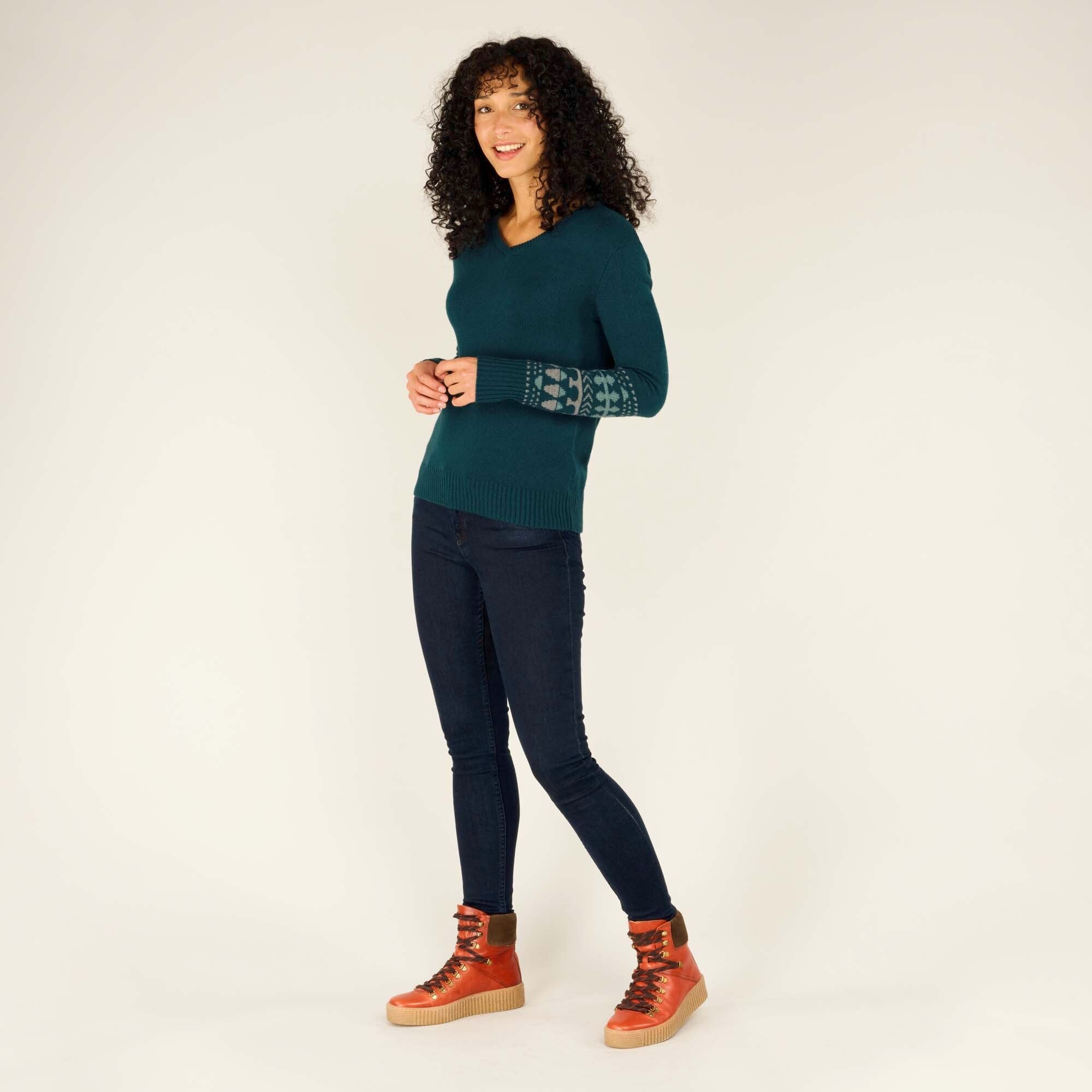 The same model stands in a relaxed pose, facing slightly to the side with her hands loosely held together in front of her. She is wearing the Sherpa Adventure Gear Maya V-Neck Sweater in Green, dark blue skinny jeans, and tan-brown high-top boots with thick soles. The sleeve design of the sweater includes a mix of geometric and traditional patterns. The background remains off-white.