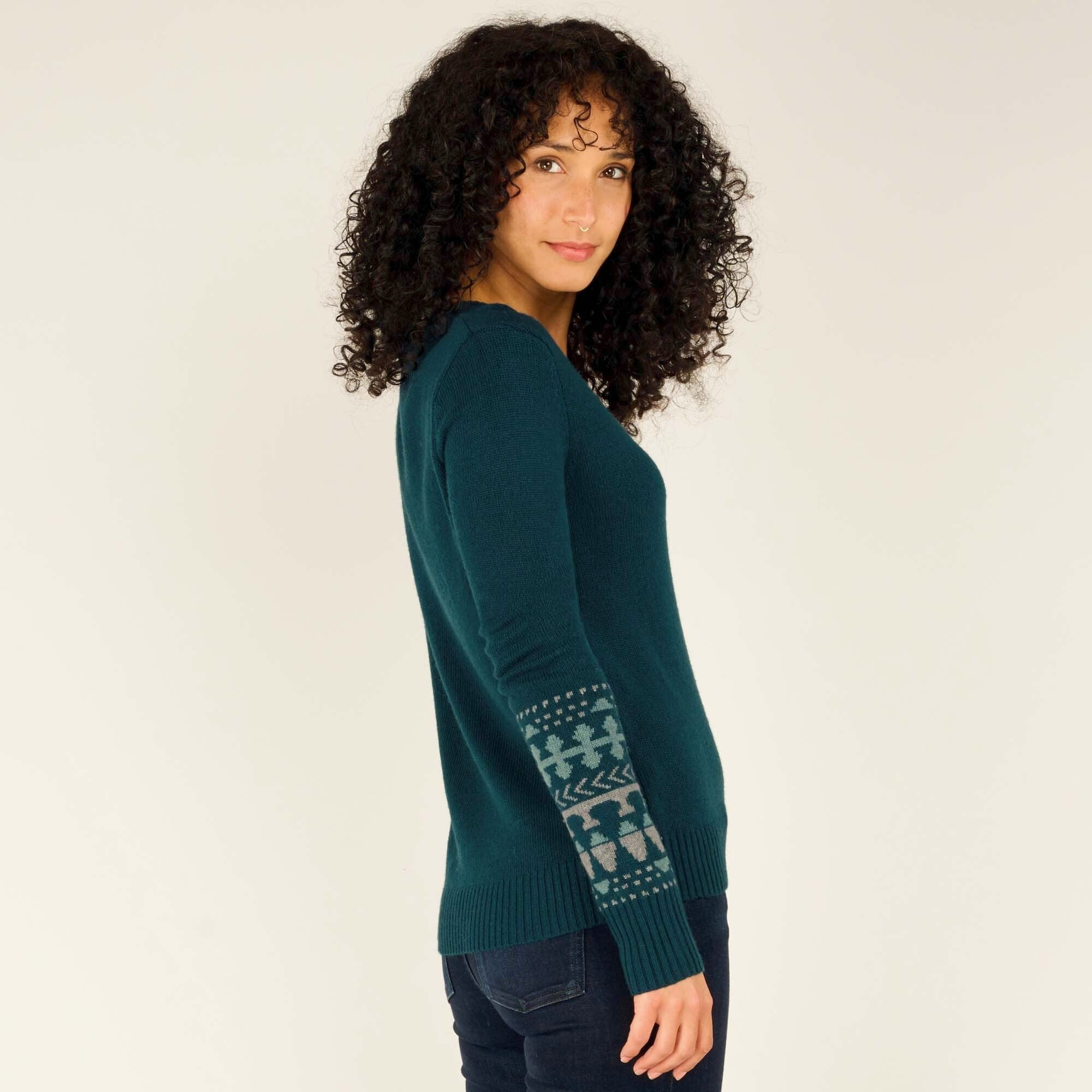 A side profile of the model as she turns slightly towards the camera with a soft expression. Her curly black hair frames her face. The Sherpa Adventure Gear Maya V-Neck Sweater in Green fits snugly, showcasing the patterned sleeve design. The ribbed hem sits just below her waist, and the dark blue jeans complement the look.