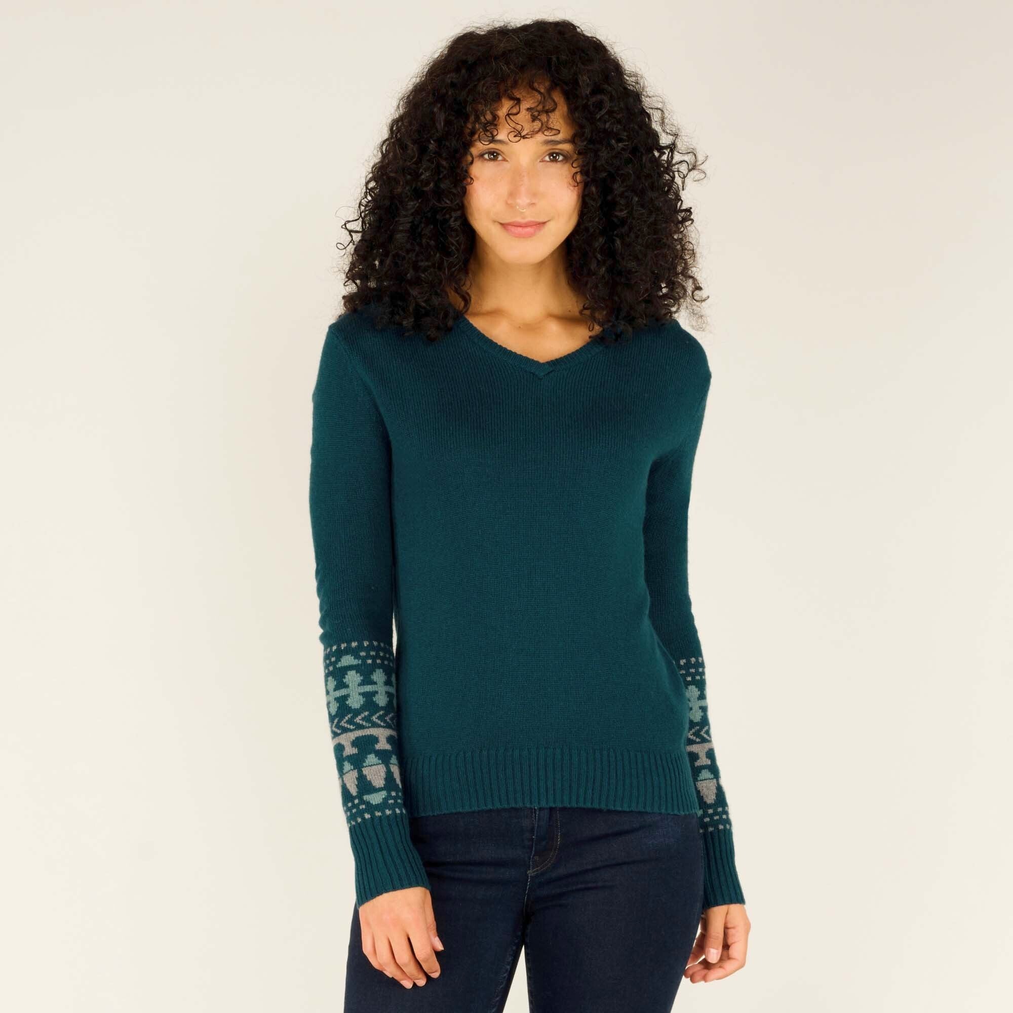 A front-facing shot of the model wearing the Sherpa Adventure Gear Maya V-Neck Sweater in Green. She has a subtle smile and gazes directly at the camera. The sweater’s fitted silhouette, intricate sleeve pattern, and soft texture are visible. The neutral background emphasizes the rich color of the sweater.