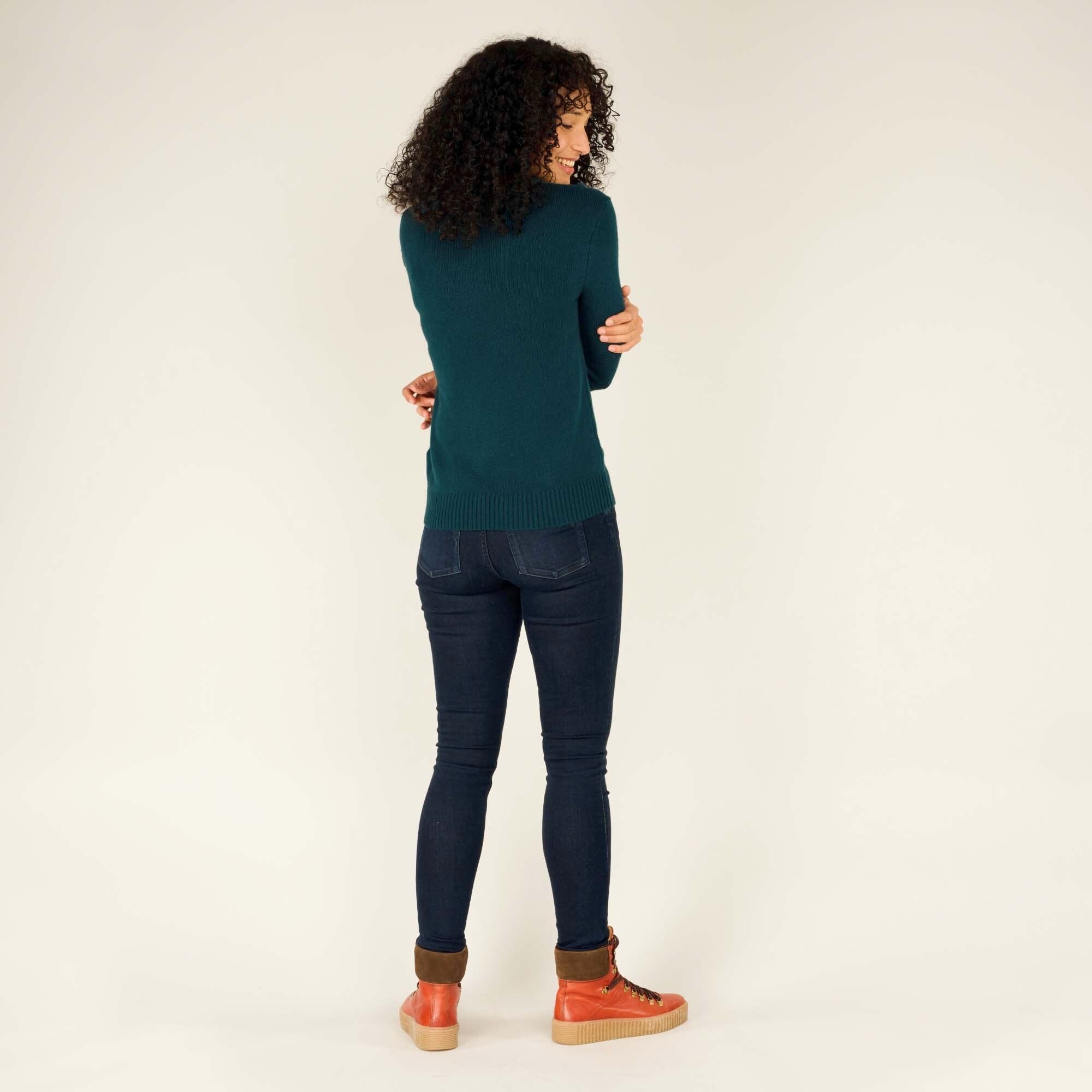 The model is shown from the back with her arms crossed over her chest, looking over her shoulder with a slight smile. The full back of the Sherpa Adventure Gear Maya V-Neck Sweater in Green is visible, along with the ribbed hem and the continuation of the sleeve pattern. Her dark blue jeans and tan-brown high-top boots complete the outfit.