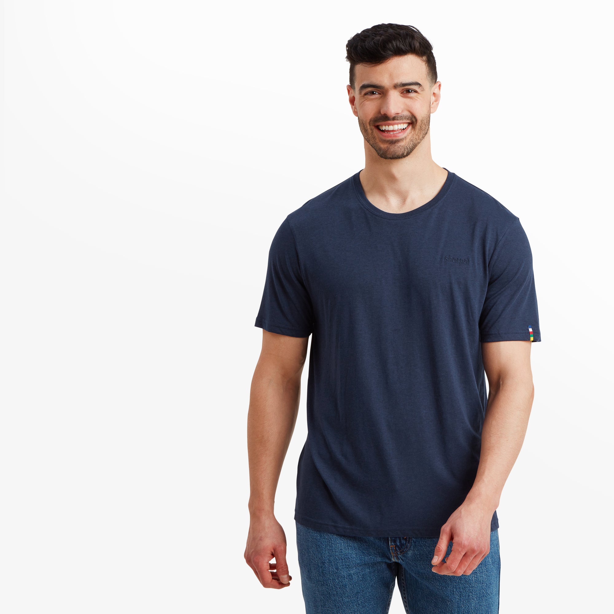 A male model stands smiling, wearing a Sherpa Adventure Gear Bali T-Shirt in Blue made of soft fabric. The shirt features a classic crew neck and short sleeves. The word "Sherpa" is subtly embroidered on the left chest in a matching navy tone. The T-shirt has a relaxed fit and pairs with casual blue jeans for a laid-back style. The model is positioned against a clean white background.