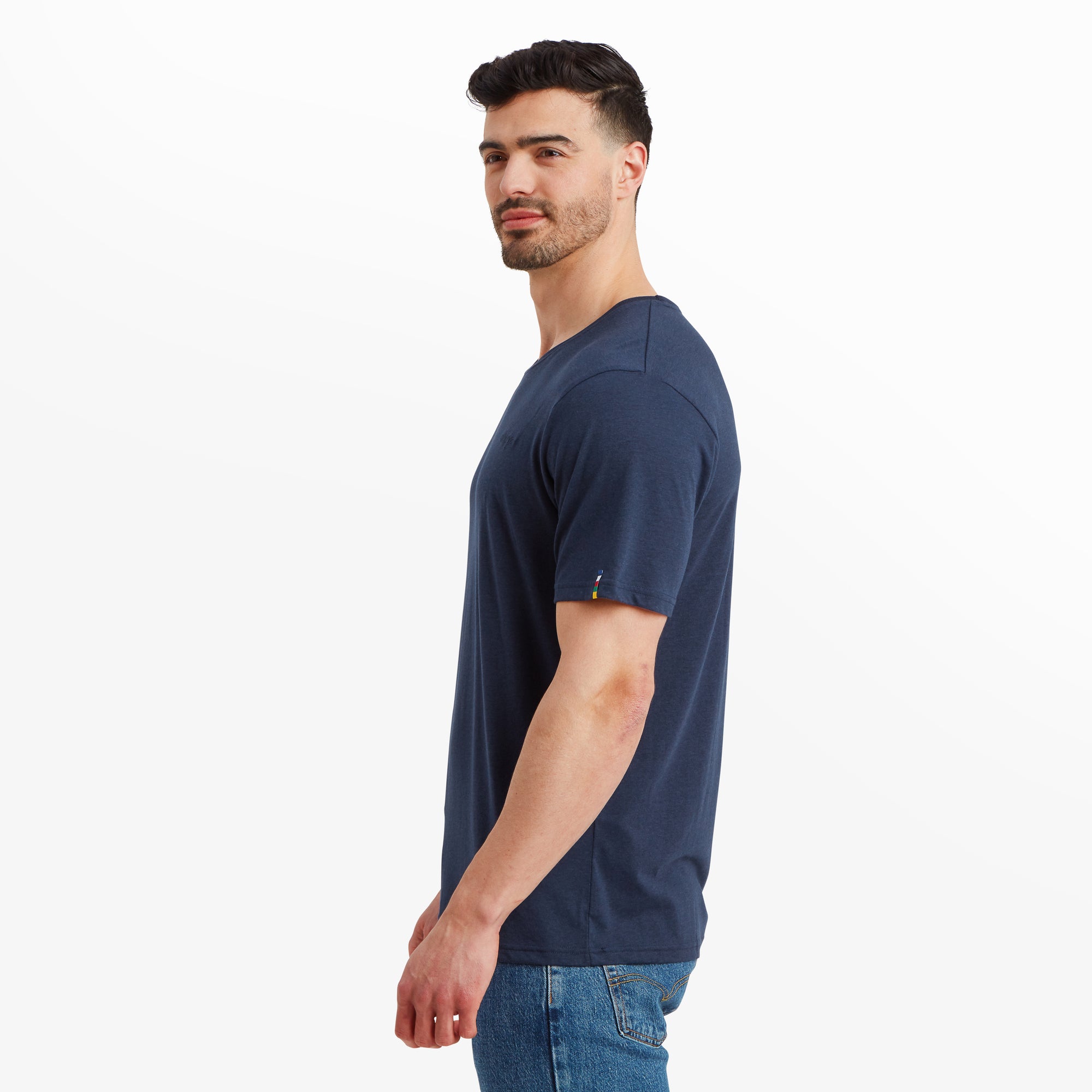 The male model turns to his side, showcasing the Sherpa Adventure Gear Bali T-Shirt in Blue’s relaxed fit. The sleeve length and stitching details are visible, emphasising the minimalist design. The hemline rests neatly on the waistband of his blue jeans. The Sherpa brand’s colorful woven flag tag is subtly visible on the left sleeve.