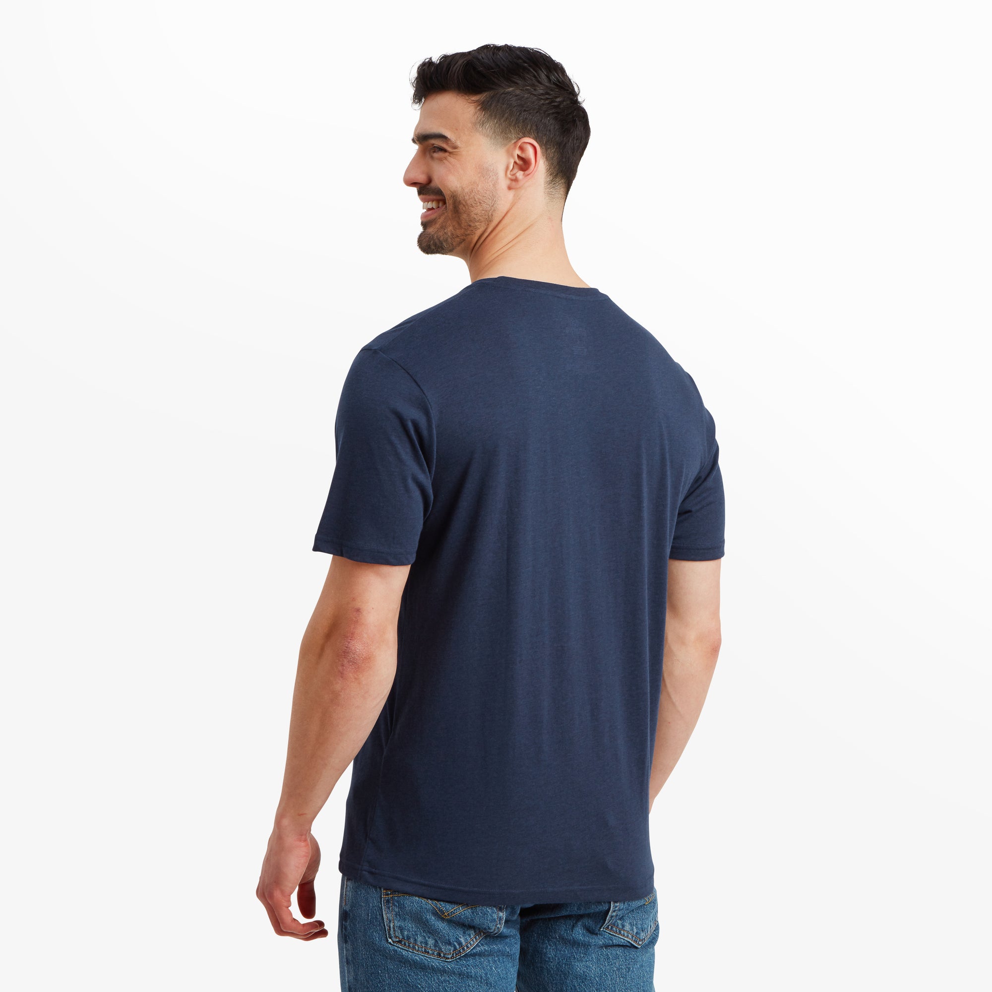 The model faces away from the camera, displaying the clean and smooth back of the Sherpa Adventure Gear Bali T-Shirt in Blue. The navy fabric flows effortlessly without any seams or designs interrupting the simplicity. The shirt rests comfortably along the shoulders and hangs naturally at the waist, complementing the casual aesthetic.