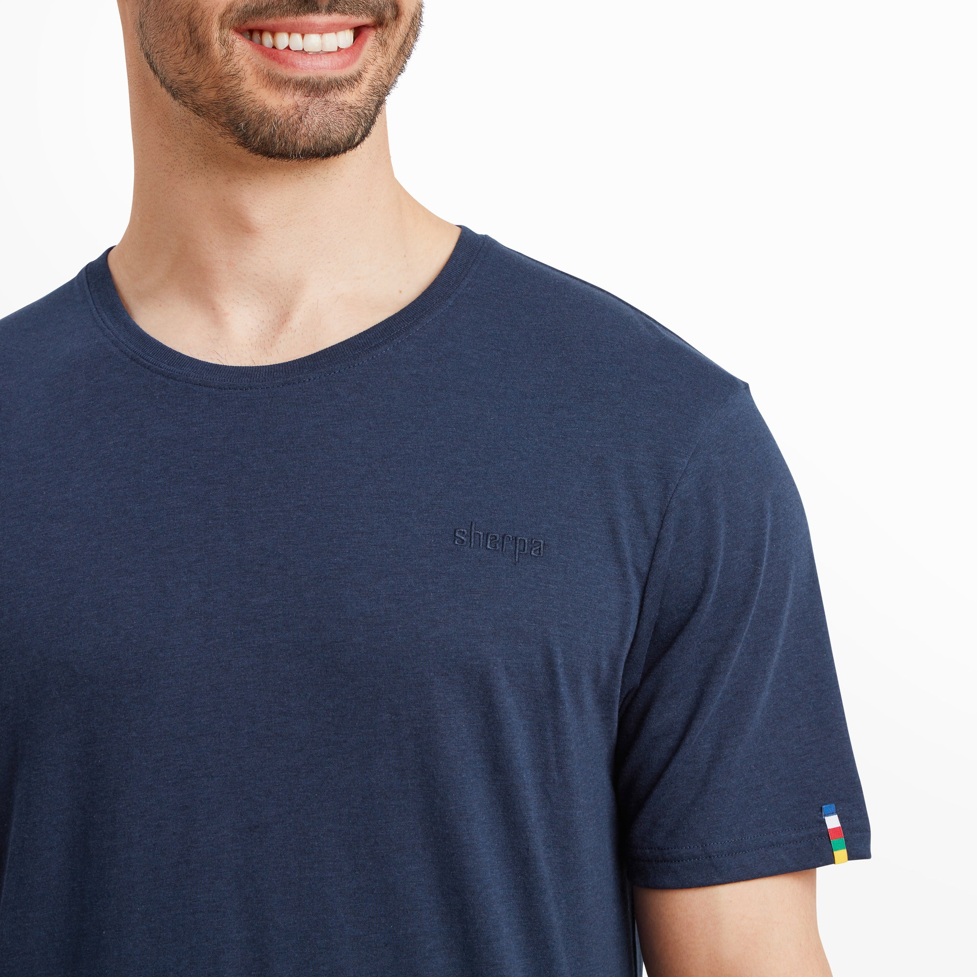 A detailed close-up shot focuses on the chest area of the Sherpa Adventure Gear Bali T-Shirt in Blue, highlighting the small embroidered "Sherpa" logo in a matching navy thread. The texture of the soft fabric and the precision of the embroidery are clearly visible. The sleeve's small colorful woven flag tag adds a subtle pop of color to the otherwise monochromatic design.