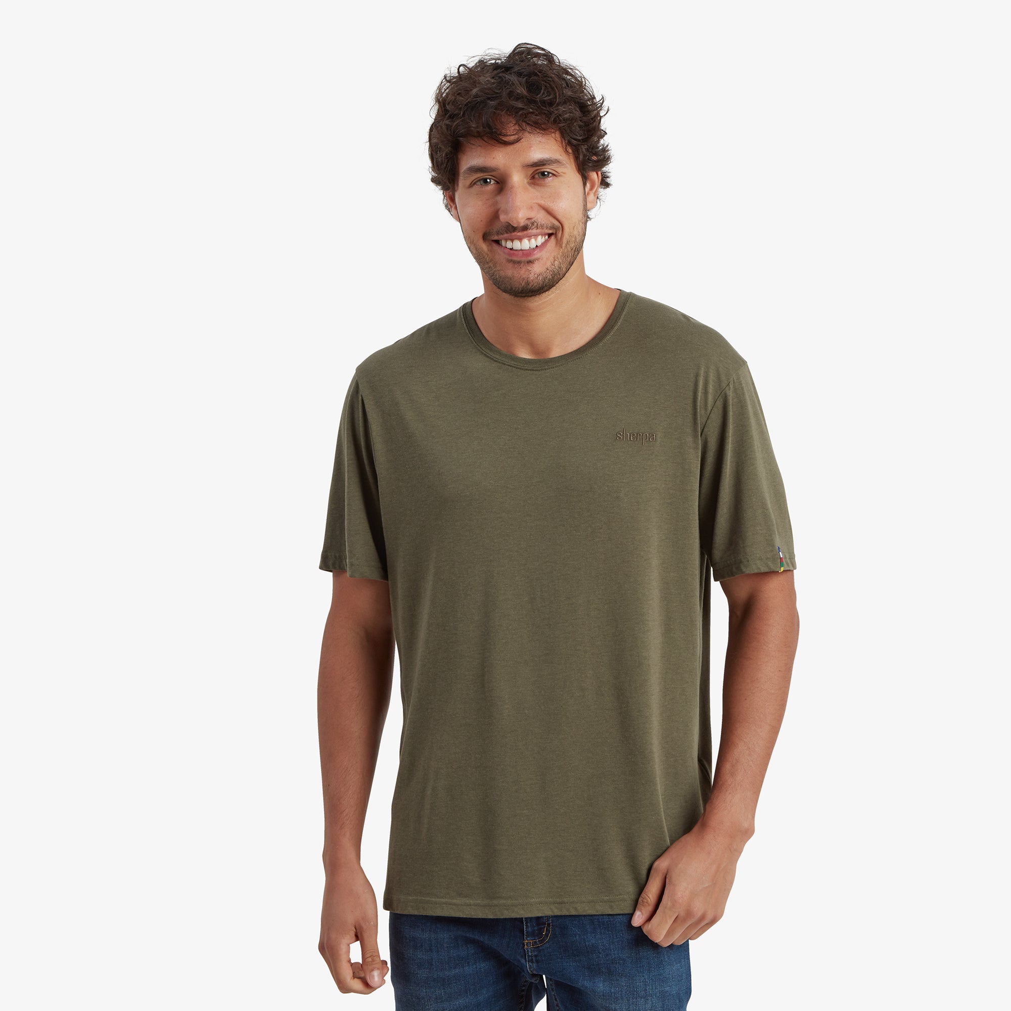 A male model is wearing a simple Sherpa Adventure Gear Bali T-Shirt in Green with a relaxed fit. The t-shirt has a round neckline and a subtle "Sherpa" logo embroidered on the left side of the chest. The model is smiling and standing in a casual pose with his hand partially in his pocket, pairing the t-shirt with blue jeans.