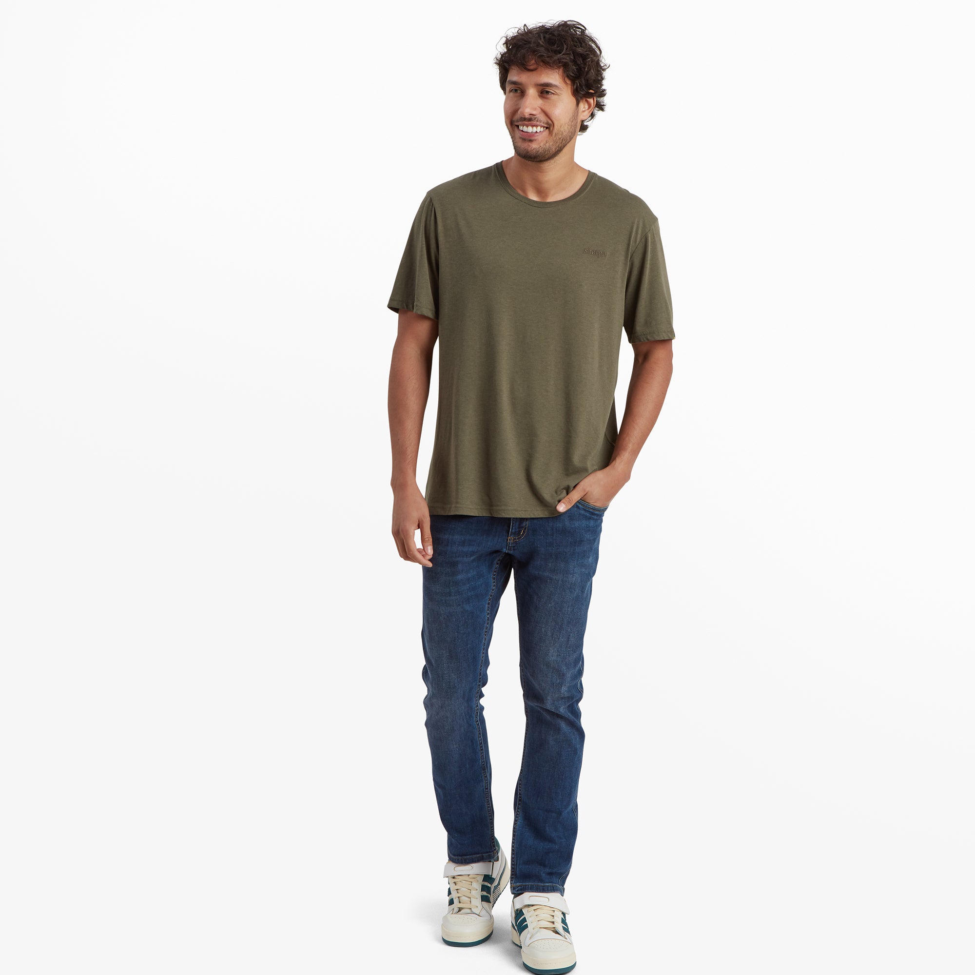 The same Sherpa Adventure Gear Bali T-Shirt in Green is shown on the male model in a full-body pose. He is standing upright, smiling, and pairing the t-shirt with slim-fit blue jeans and trainers. The t-shirt’s relaxed cut and casual style are highlighted in this pose.