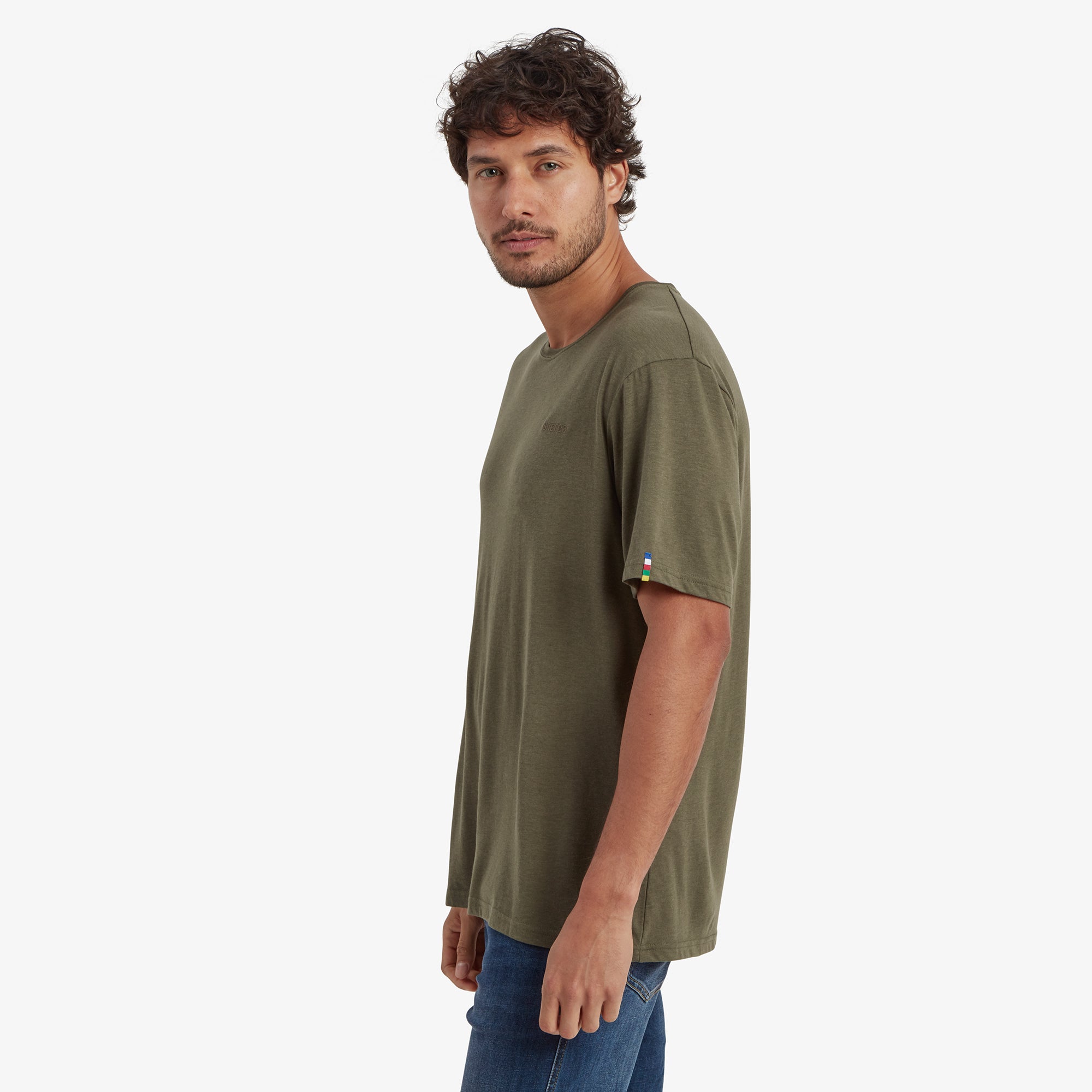 The model is turned to his left, showcasing the side profile of the Sherpa Adventure Gear Bali T-Shirt in Green. The sleeve length and a colorful fabric tag featuring horizontal stripes in red, blue, yellow, green, and white are visible on the edge of the left sleeve.