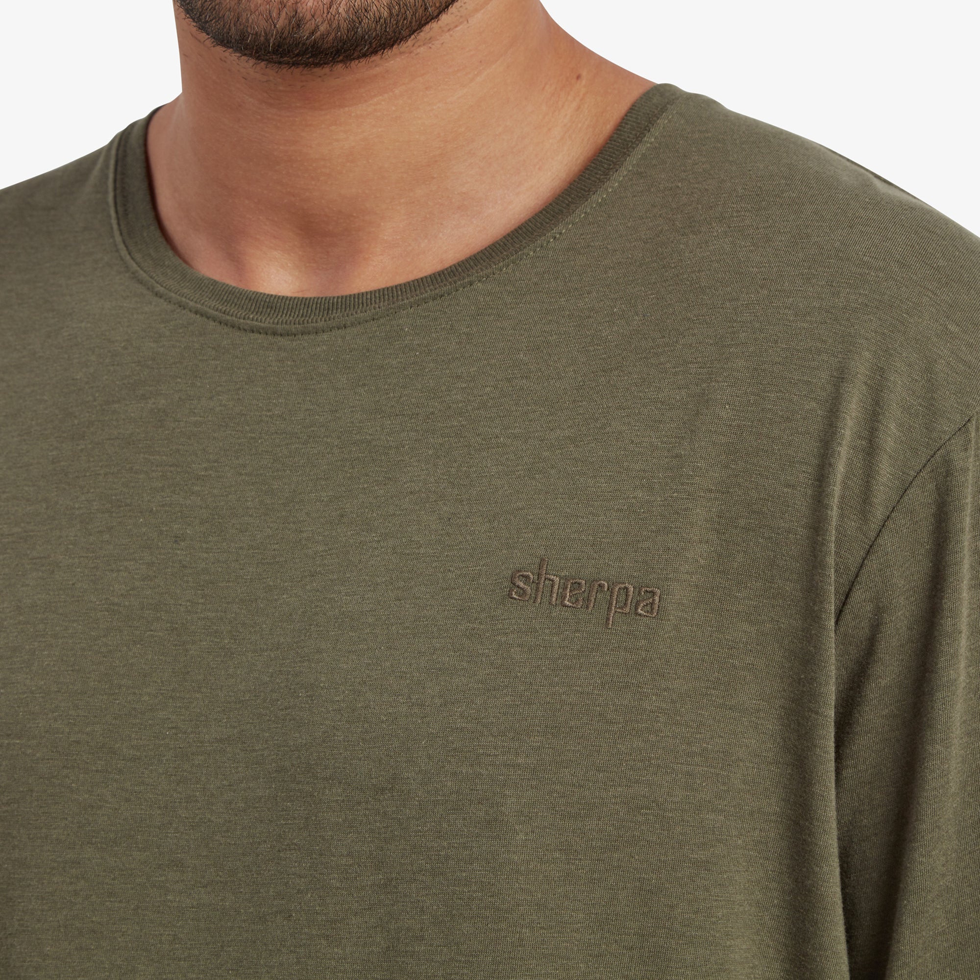 A zoomed-in shot of the "Sherpa" logo, embroidered in a tonal shade of green that blends subtly with the fabric on the Sherpa Adventure Gear Bali T-Shirt in Green. The neckline stitching is also visible, showcasing the quality of the construction.