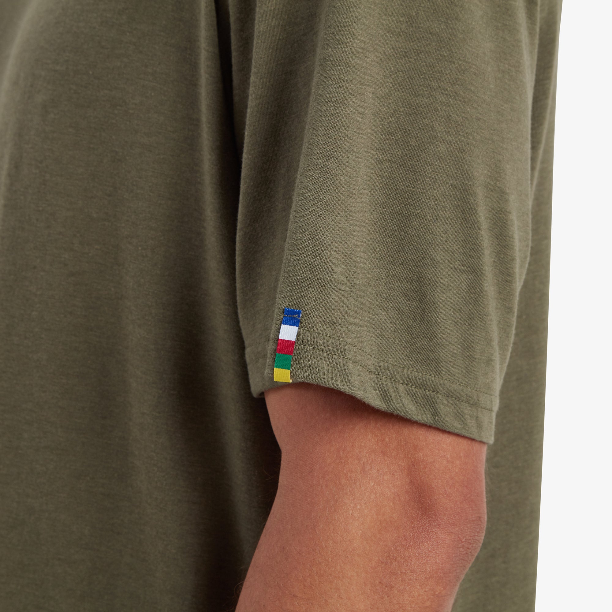 A detailed shot of the colorful fabric tag stitched onto the edge of the sleeve. The tag features a series of horizontal stripes in bold colors—red, blue, yellow, green, and white—adding a vibrant accent to the Sherpa Adventure Gear Bali T-Shirt in Green's understated design. The texture of the fabric is also visible in this close-up.