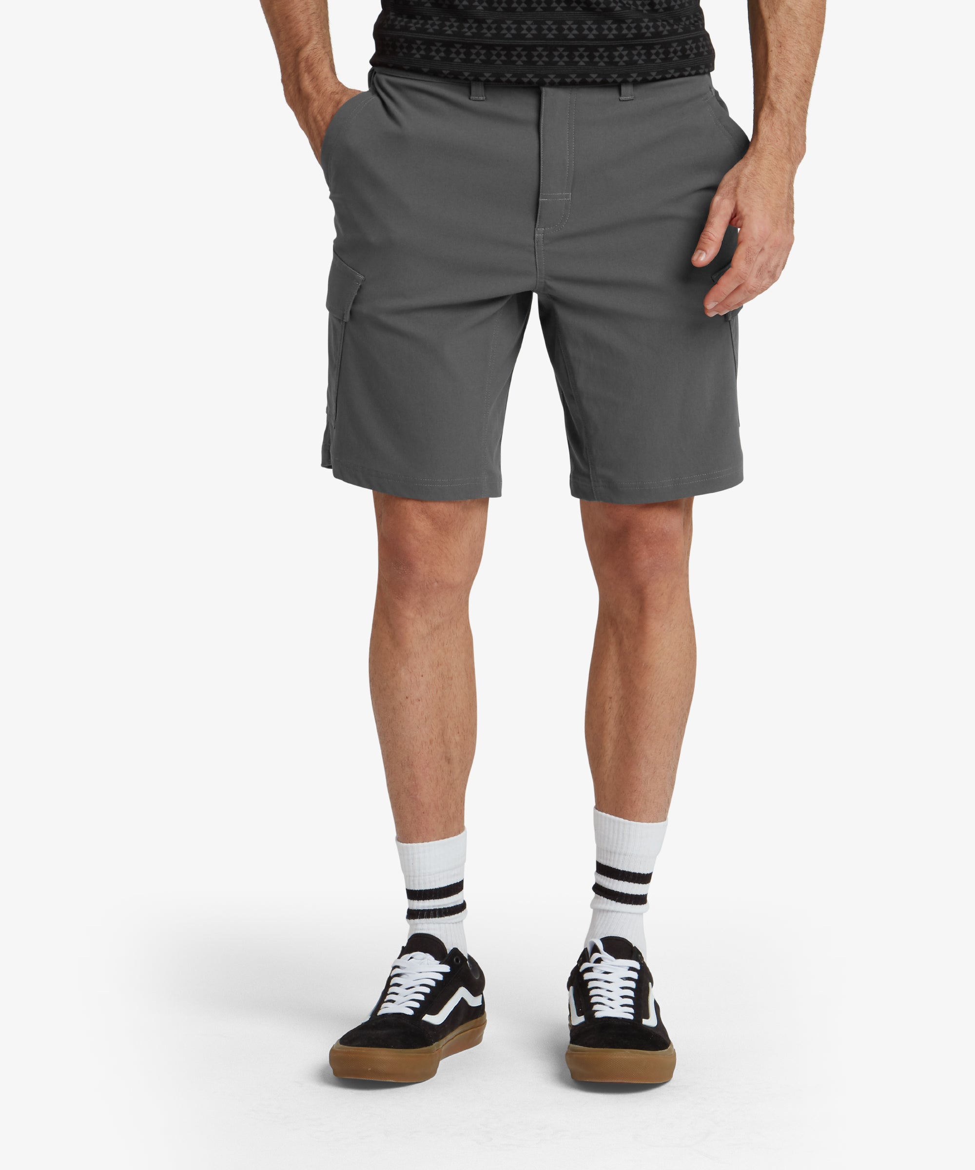 A front view of a pair of Sherpa Adventure Gear Bara Cargo Shorts in Grey worn by a model. The shorts feature two spacious cargo pockets on either side and standard side pockets. They are paired with a tucked-in olive green button-up shirt and white trainers, showcasing a clean and practical outdoor style.