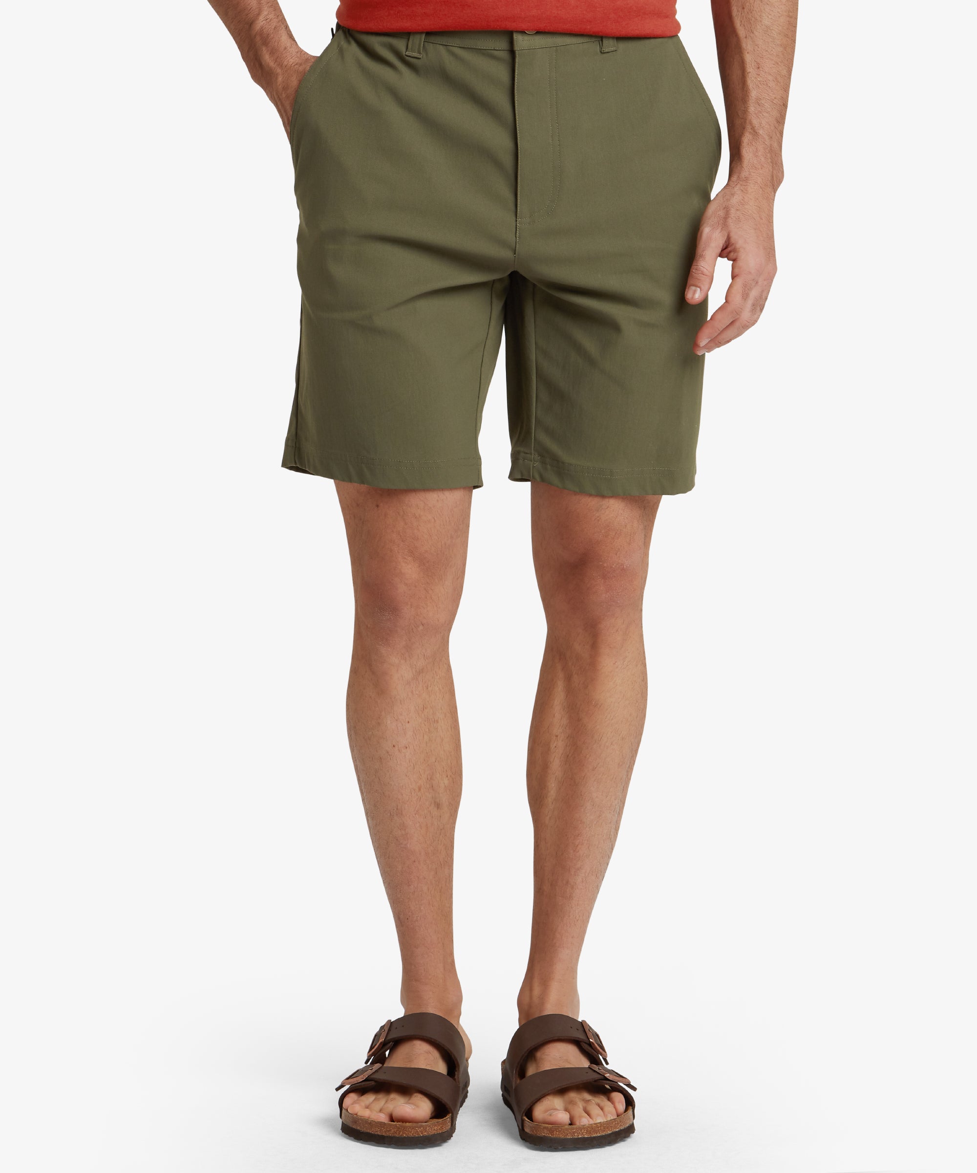 A frontal view of a model wearing a pair of Sherpa Adventure Gear Bara Shorts in Green with a clean and minimal design. The shorts have a tailored fit with belt loops, a button closure, and a zipper fly. The model pairs the shorts with white trainers, emphasising a casual and versatile style.