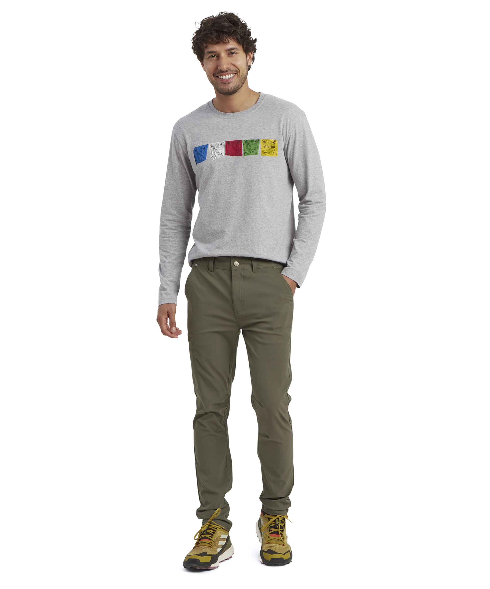 A model smiling while wearing the Sherpa Adventure Gear Bara trousers in Green with a gray graphic long-sleeve t-shirt. The full-body image showcases the trousers' streamlined fit and the casual styling paired with colorful hiking shoes.