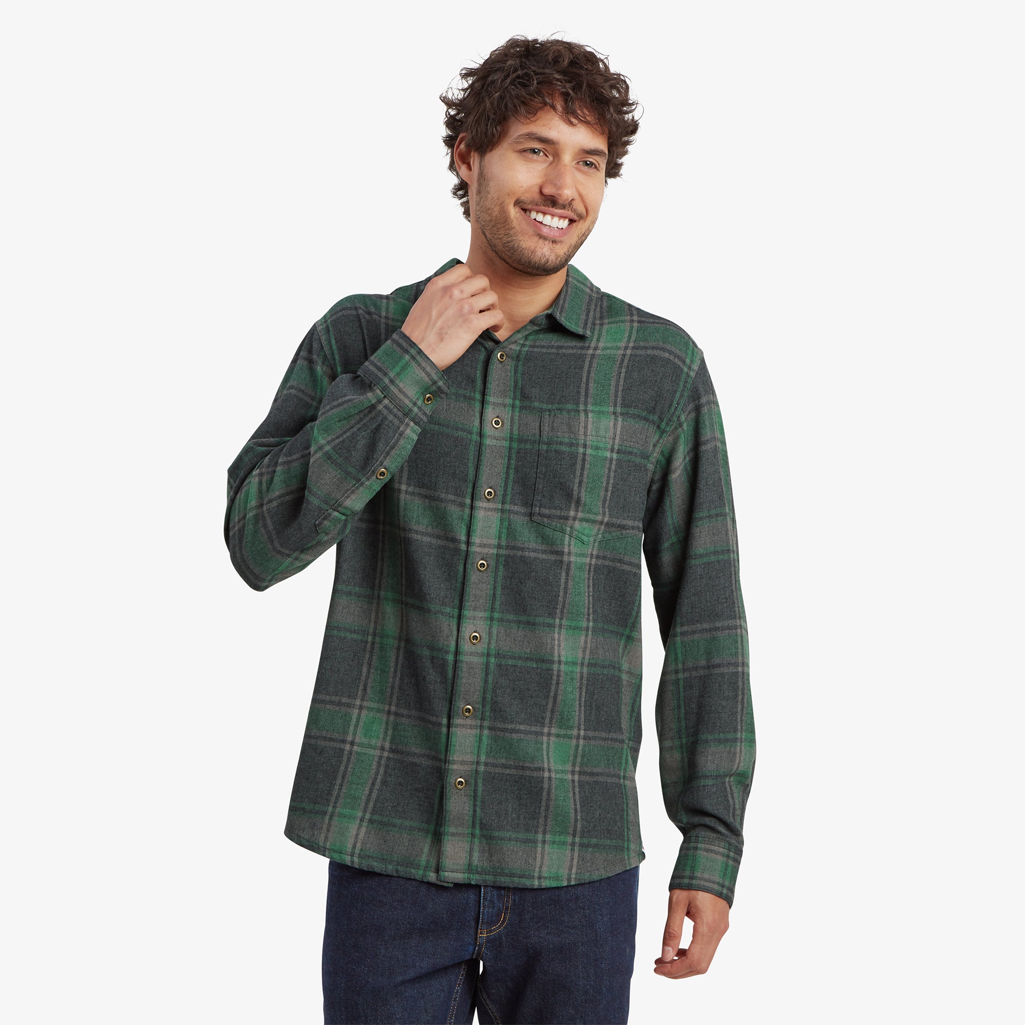 A man wearing a Sherpa Adventure Gear Batsa Long Sleeve Shirt in Black with a single chest pocket, standing with one hand near his collar and a slight smile. The shirt is buttoned up and paired with dark blue jeans, creating a casual yet neat appearance.