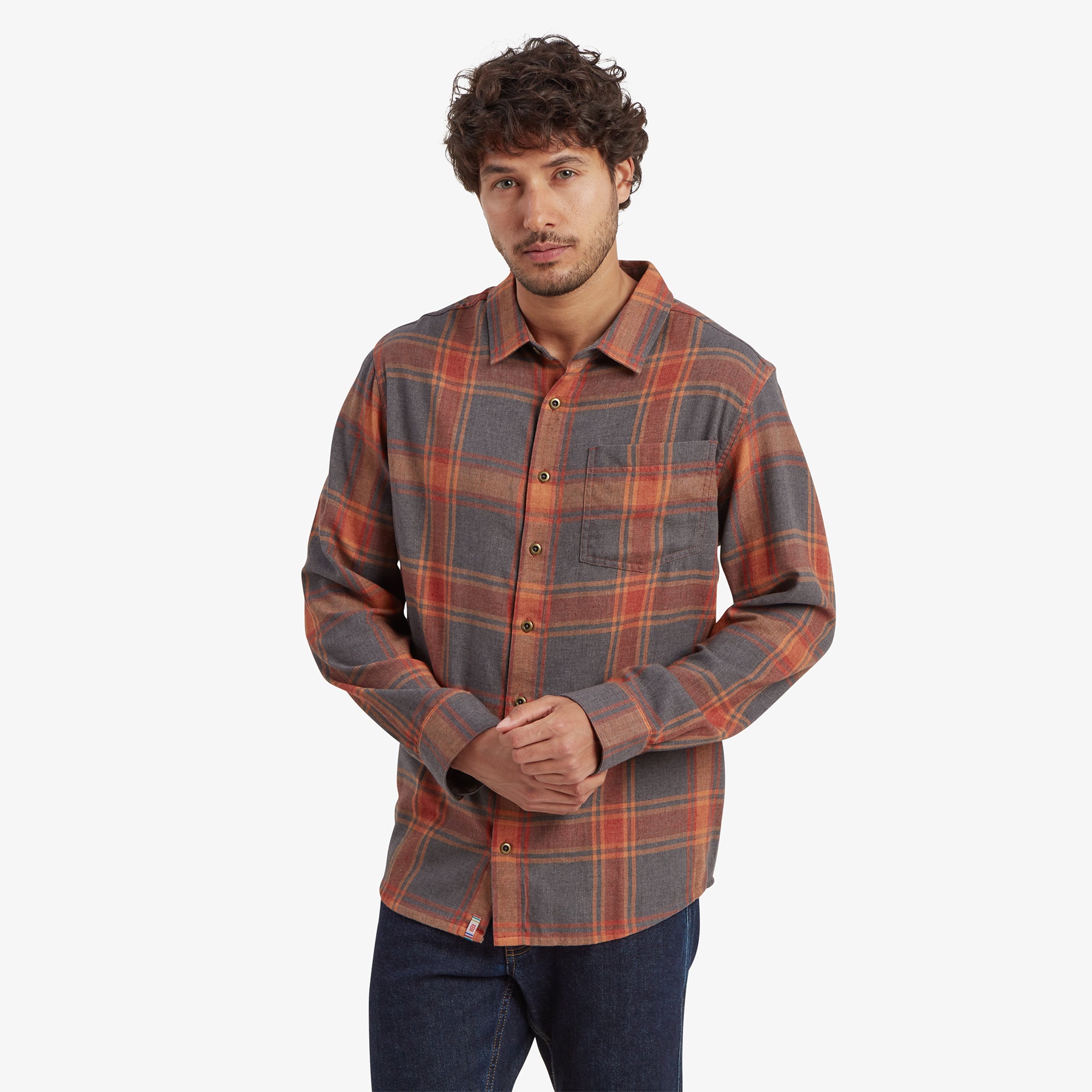 A man is wearing a Sherpa Adventure Gear Batsa Long Sleeve Shirt in Red with a button-up design. The shirt features a mix of red, orange, and gray tones with a classic chest pocket. The model is standing with his hands together, displaying the shirt's clean, tailored fit.