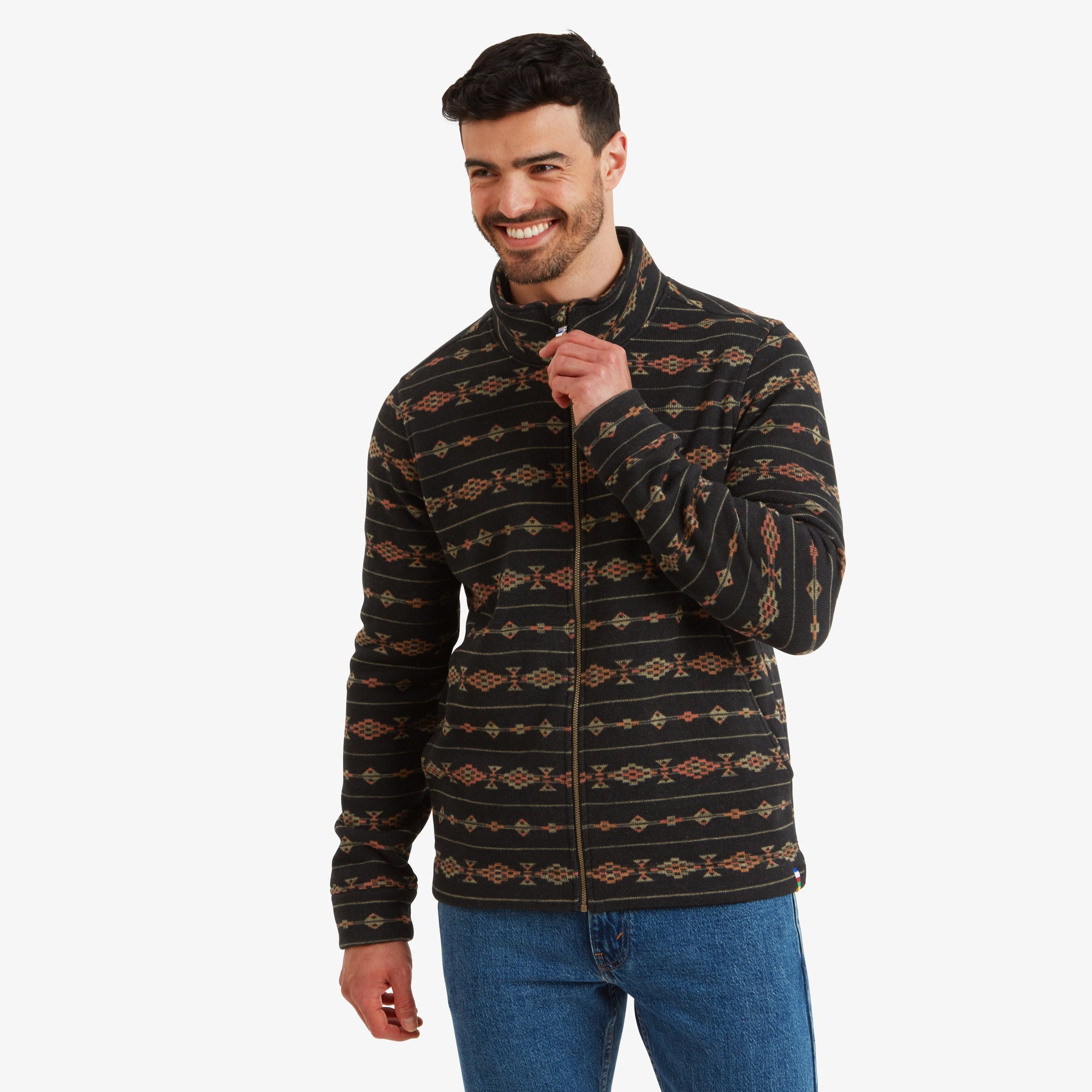 The model is wearing a Sherpa Adventure Gear Bhutan Full Zip Jacket in Black with intricate orange and yellow geometric patterns across the fabric. The front view highlights the high collar and zipper, with the model smiling and partially zipping up the jacket for a casual and approachable look.