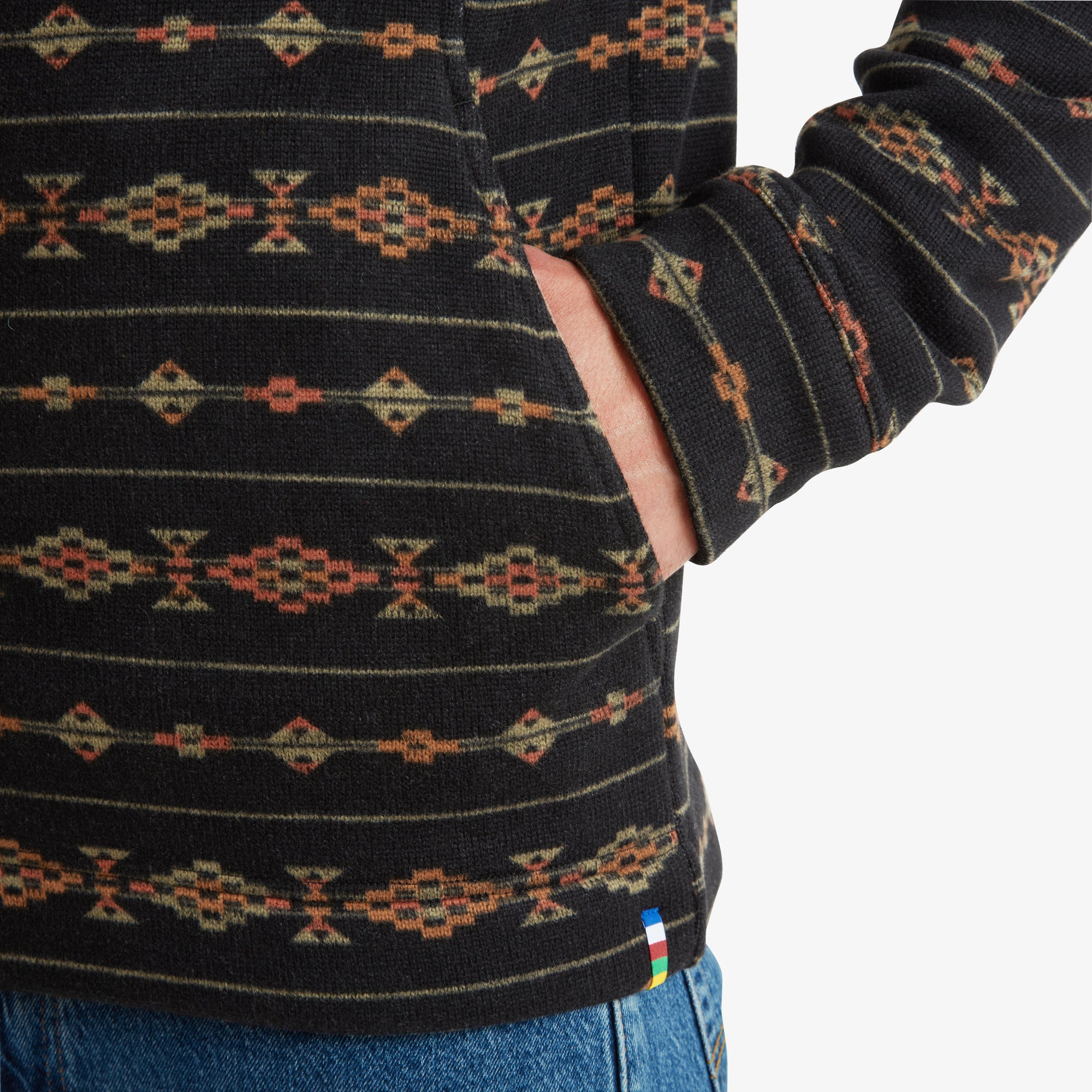 This detailed image highlights the Sherpa Adventure Gear Bhutan Full Zip Jacket in Black's pocket and the seamless integration of the design. The pocket is functional yet discreet, blending perfectly into the geometric pattern. The Sherpa tag with multicolored stripes is visible at the hem, adding a pop of color.