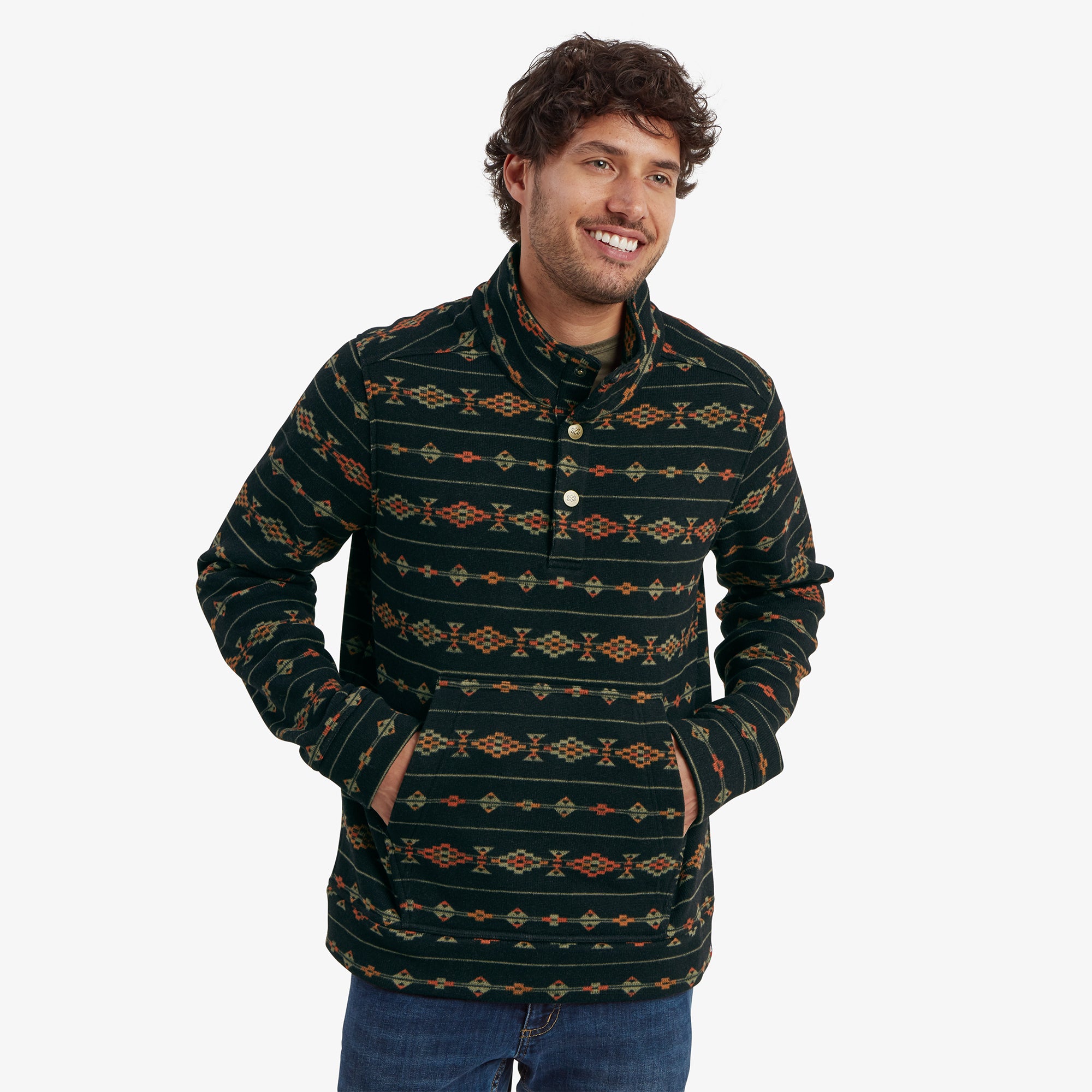 A smiling man wearing a Sherpa Adventure Gear Bhutan Pullover in Black with intricate, colorful geometric patterns. The pullover features a high collar with three decorative buttons and a front kangaroo pocket. The man has his hands in the pocket and is posed casually.