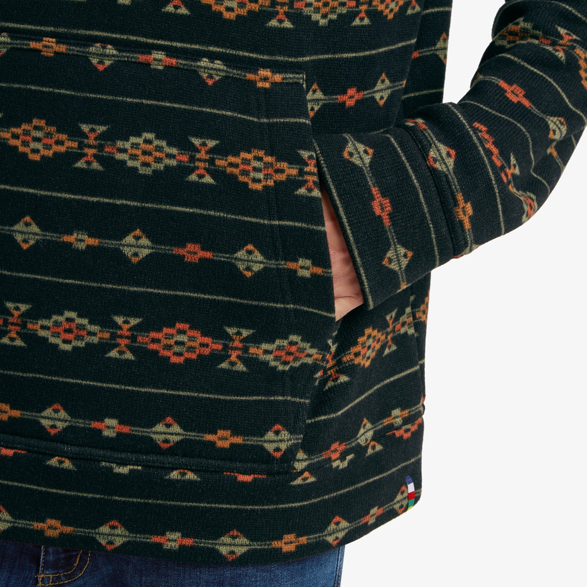 A detailed close-up of the kangaroo pocket of the Sherpa Adventure Gear Bhutan Pullover in Black. The colorful geometric design continues seamlessly, and the man's hand is partially inserted into the pocket. The edge of a small branded flag tag is visible near the pocket's seam.