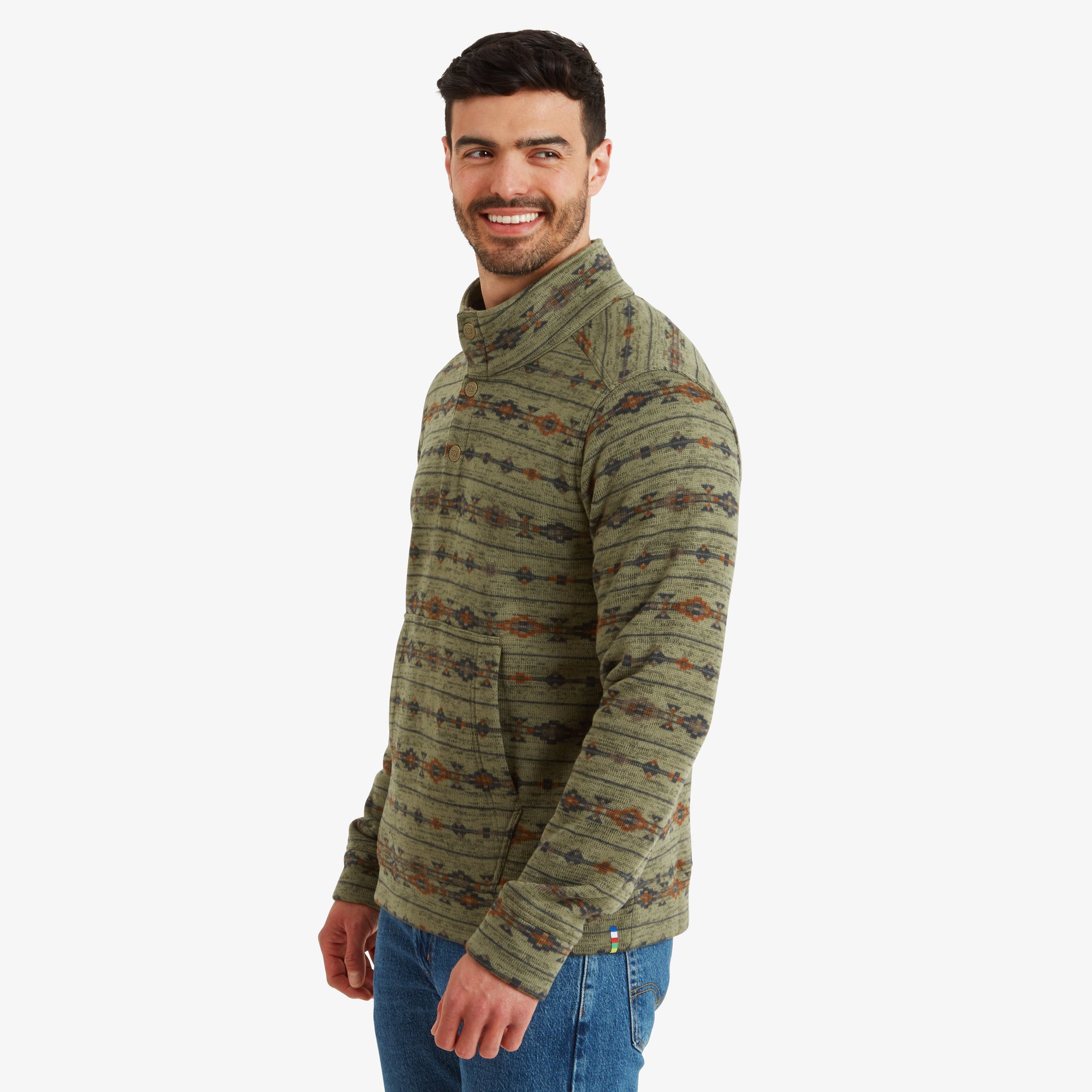 A side profile shot highlights the detailed stitching along the shoulder and sleeves of the Sherpa Adventure Gear Bhutan Pullover in Green. The man turns slightly to smile at the camera, showcasing the fit and style of the garment. The green, orange, and navy tones of the fabric create a warm and earthy feel.