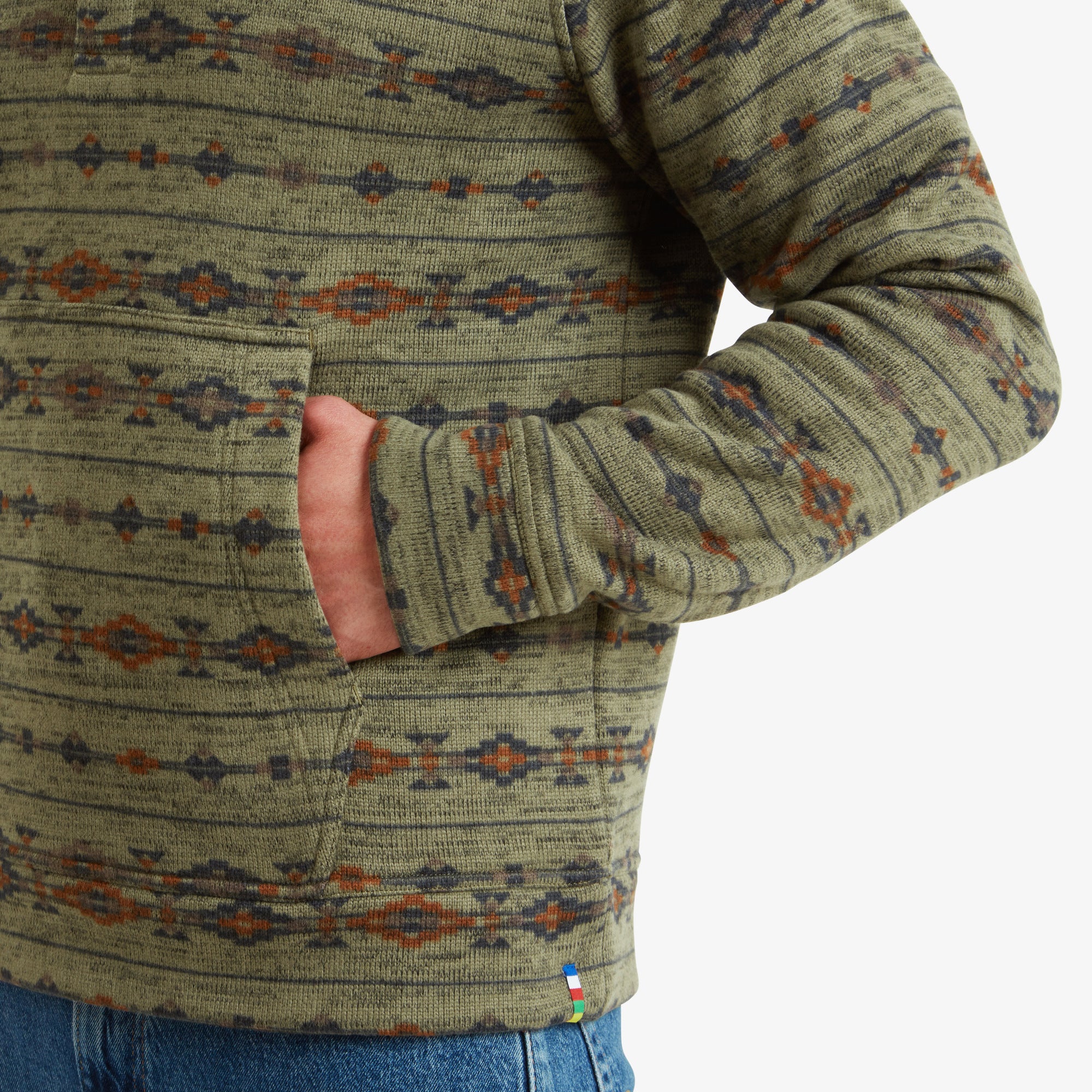 A detailed shot of the front pocket, with the man’s hand partially inside it. The pocket is seamlessly integrated into the design, and the image highlights the textured fabric of the Sherpa Adventure Gear Bhutan Pullover in Green. A small multicolor stitched tab is visible near the hem, adding a subtle branding detail.