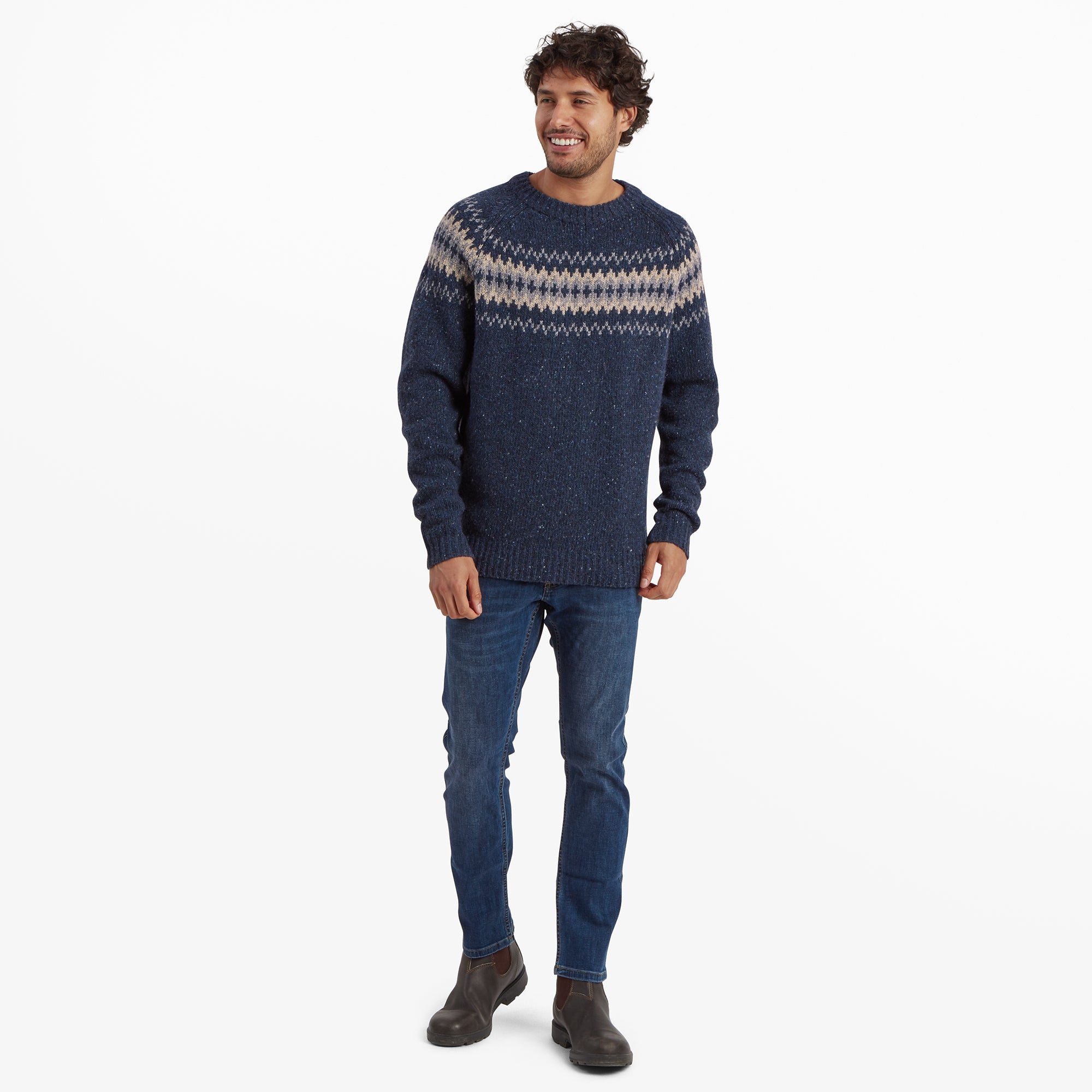 The same man is captured in a full-body pose, wearing the Sherpa Adventure Gear Dumji Sweater in Blue paired with fitted blue jeans and dark brown boots. He is smiling and standing confidently, giving a full view of the sweater's length and overall style.