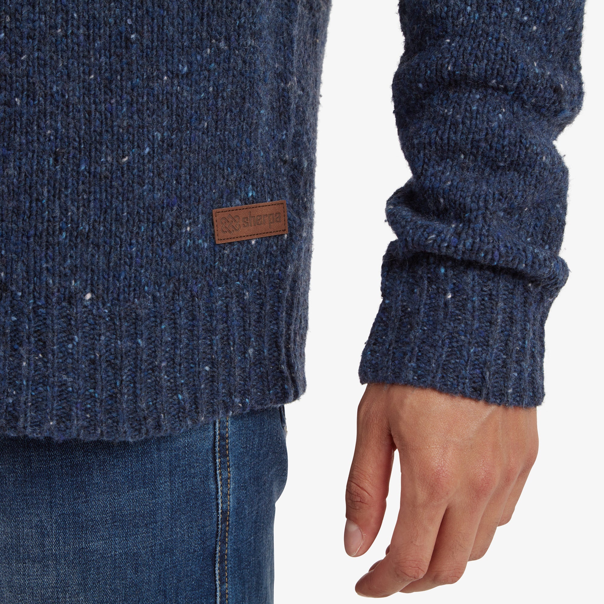 A focused shot of the ribbed hem and cuff at the wrist on the Sherpa Adventure Gear Dumji Sweater in Blue, showing a close view of the texture and a small leather Sherpa brand tag sewn into the lower edge of the sweater. The textured fabric adds a rustic, hand-crafted appeal.