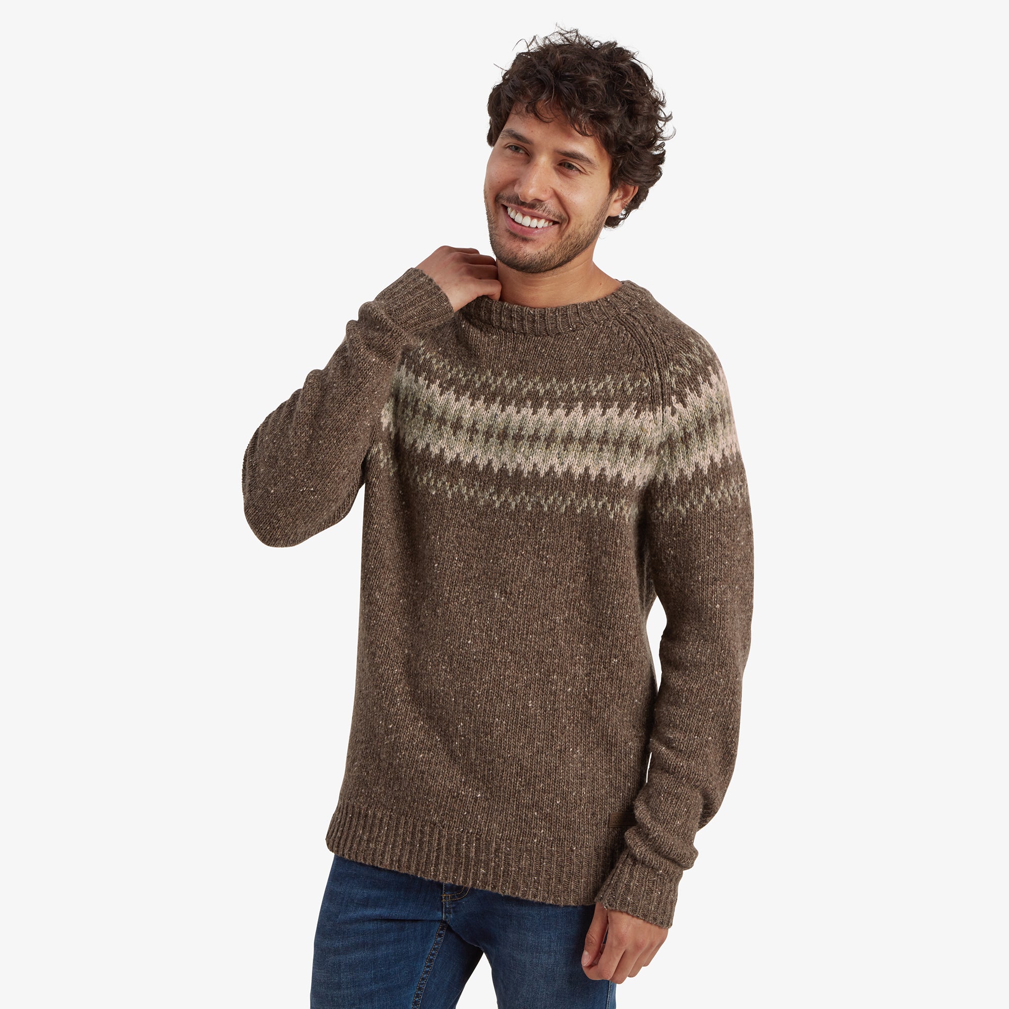 A male model with curly dark hair and light facial stubble wears a Sherpa Adventure Gear Dumji Sweater in Brown with a traditional Fair Isle pattern across the chest in muted green and cream tones. The sweater has a ribbed crew neckline, cuffs, and hem. He is smiling and lightly touching his neck with his right hand, looking relaxed. He pairs the sweater with medium-wash blue jeans against a plain white background.