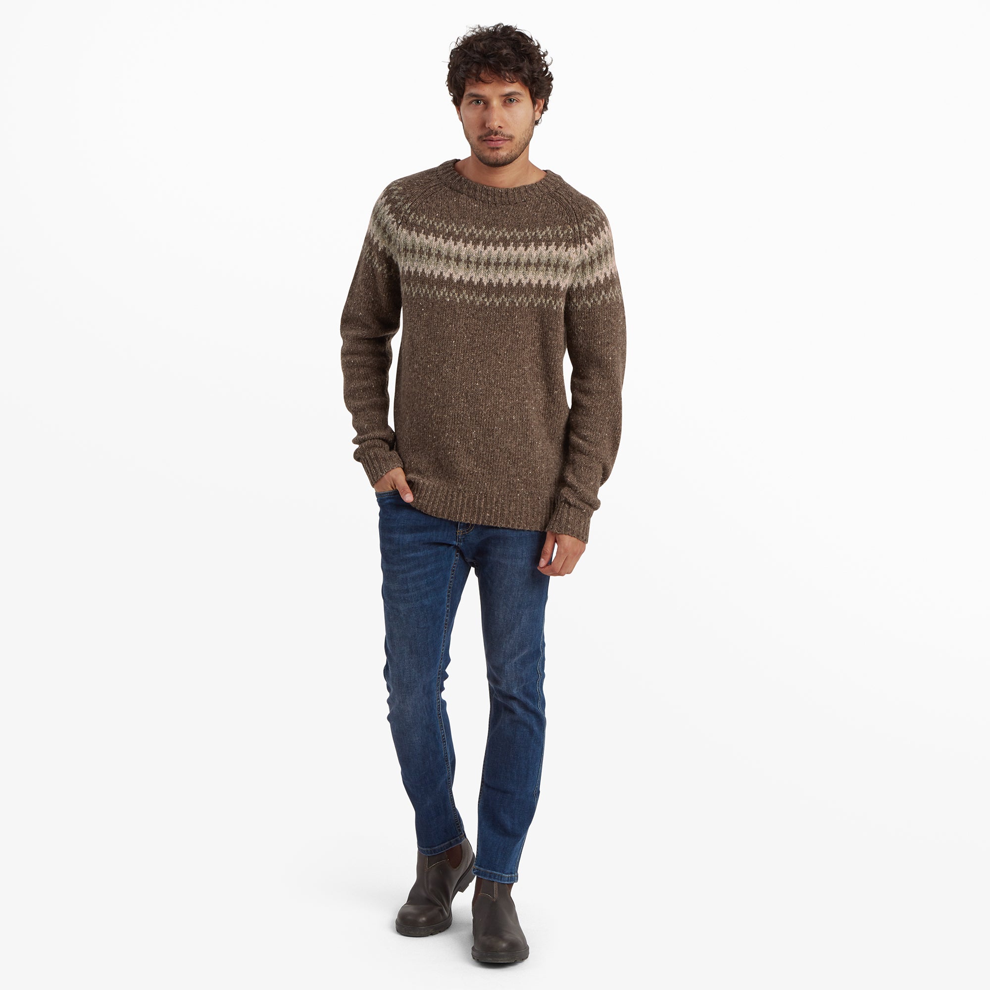 The model is shown in a full-body pose wearing the Sherpa Adventure Gear Dumji Sweater in Brown and blue jeans. He pairs the look with dark brown leather boots. His hands are relaxed at his sides as he stands slightly angled toward the camera, looking confident and composed.