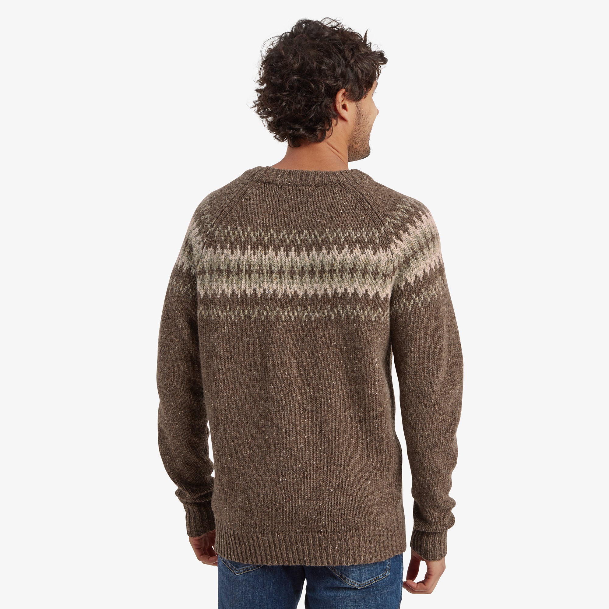 The back view of the model wearing the Sherpa Adventure Gear Dumji Sweater in Brown, highlighting the continuation of the Fair Isle pattern across the upper back. The ribbed collar and cuffs are visible, and the sweater has a relaxed fit. His curly hair adds texture to the scene as he looks away from the camera.