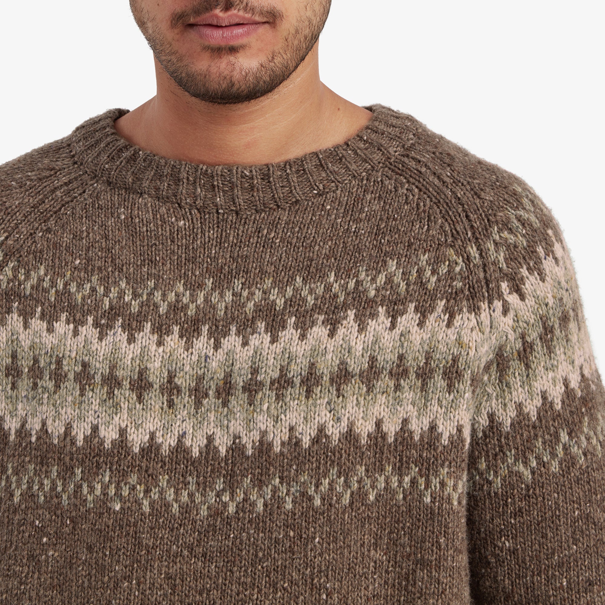 A close-up of the Sherpa Adventure Gear Dumji Sweater in Brown’s chest area, displaying the intricate Fair Isle knit pattern in detail. The fine texture of the wool blend material is visible, with speckled fibers adding depth to the brown base color. The ribbed crew neckline is also clearly defined.