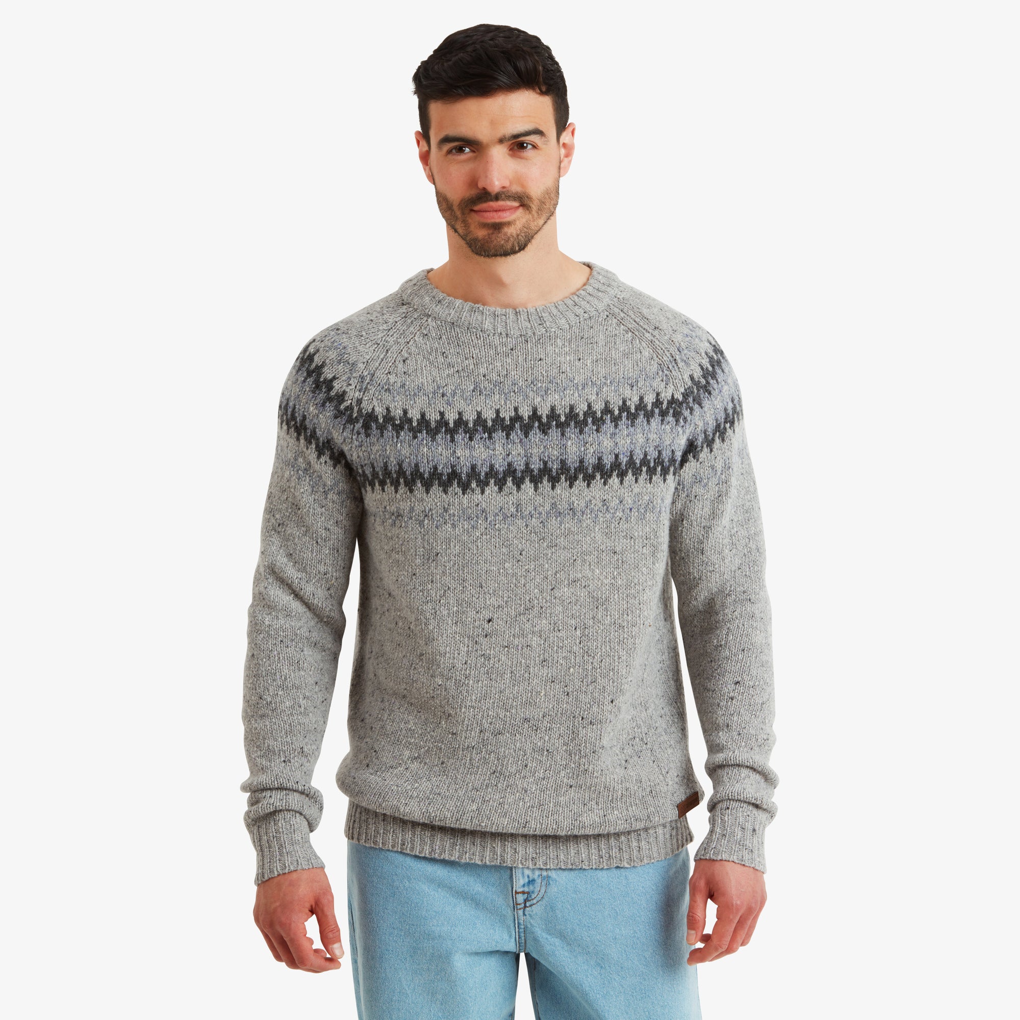 A model wearing a Sherpa Adventure Gear Dumji Sweater in Brown with a round neck, featuring a traditional pattern in dark grey and black across the chest. The textured material highlights the cozy, hand-crafted feel of the garment. The model pairs the sweater with light-washed jeans, standing confidently facing forward.