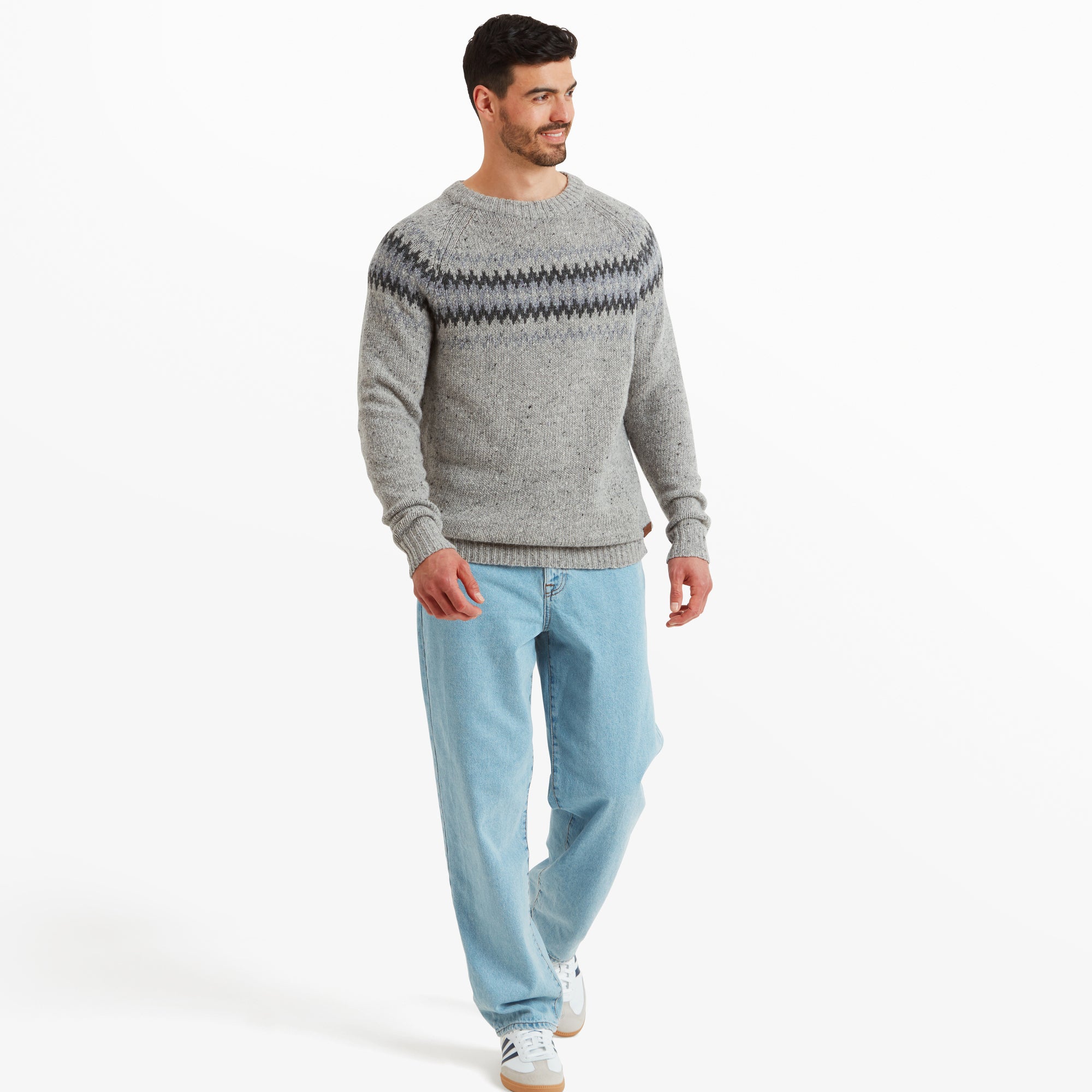 The same model in the Sherpa Adventure Gear Dumji Sweater in Brown, paired with relaxed-fit light blue jeans and white trainers. The photo is a full-body shot, showcasing the casual, comfortable style of the outfit. The model is mid-step, smiling slightly, creating a sense of movement.