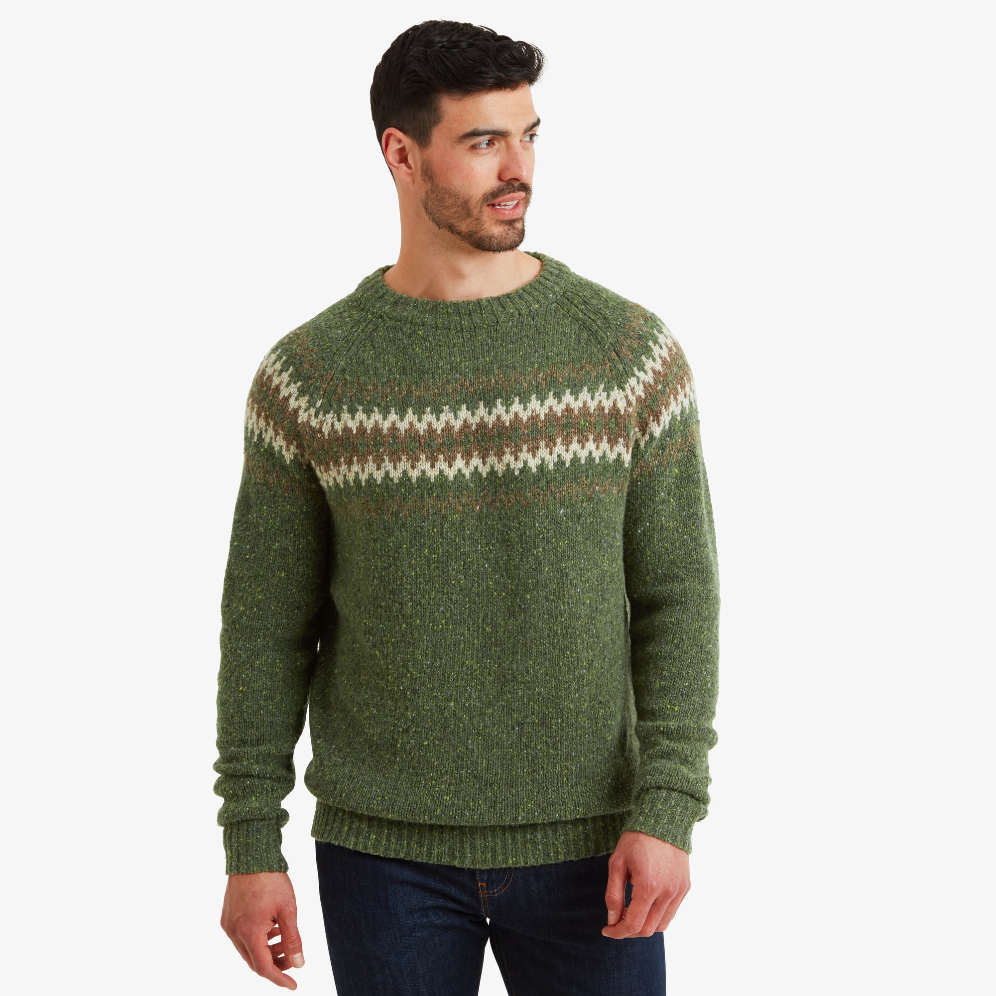 A man models a Sherpa Adventure Gear Dumji Sweater in Green featuring a fair isle pattern across the chest in white and brown tones. He looks to the side with a casual expression, showcasing the sweater's relaxed fit and soft texture. The jeans he wears complement the sweater's earthy tones.