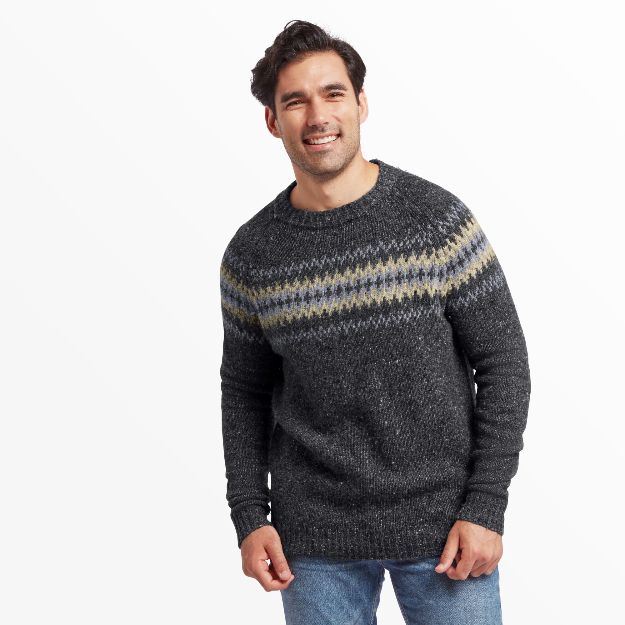 A male model with short dark hair and a smile is wearing a Sherpa Adventure Gear Dumji Sweater in Grey with a traditional Fair Isle-inspired pattern across the chest and upper arms in shades of light grey, cream, and muted olive green. The sweater has a classic crew neck and ribbed cuffs. He is standing facing the camera with his hands at his sides, paired with light blue jeans. The background is plain white.