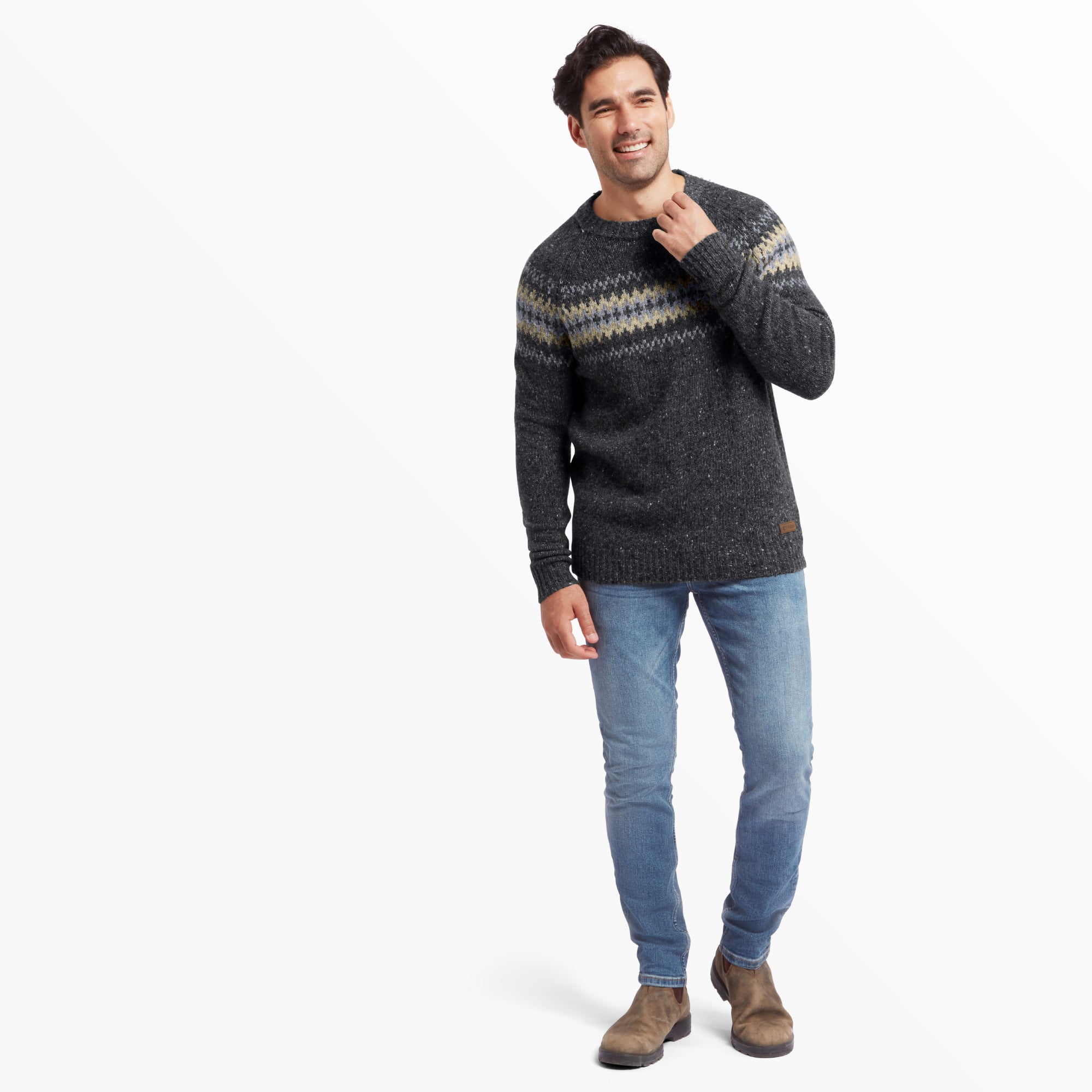 The same male model is captured in a full-body shot, wearing the Sherpa Adventure Gear Dumji Sweater in Grey with light blue jeans and brown suede boots. He is slightly turned to his right, holding the sweater collar with his right hand and smiling. The pattern across the chest is clearly visible, and the hem of the sweater falls just below his waist. The background is plain white.