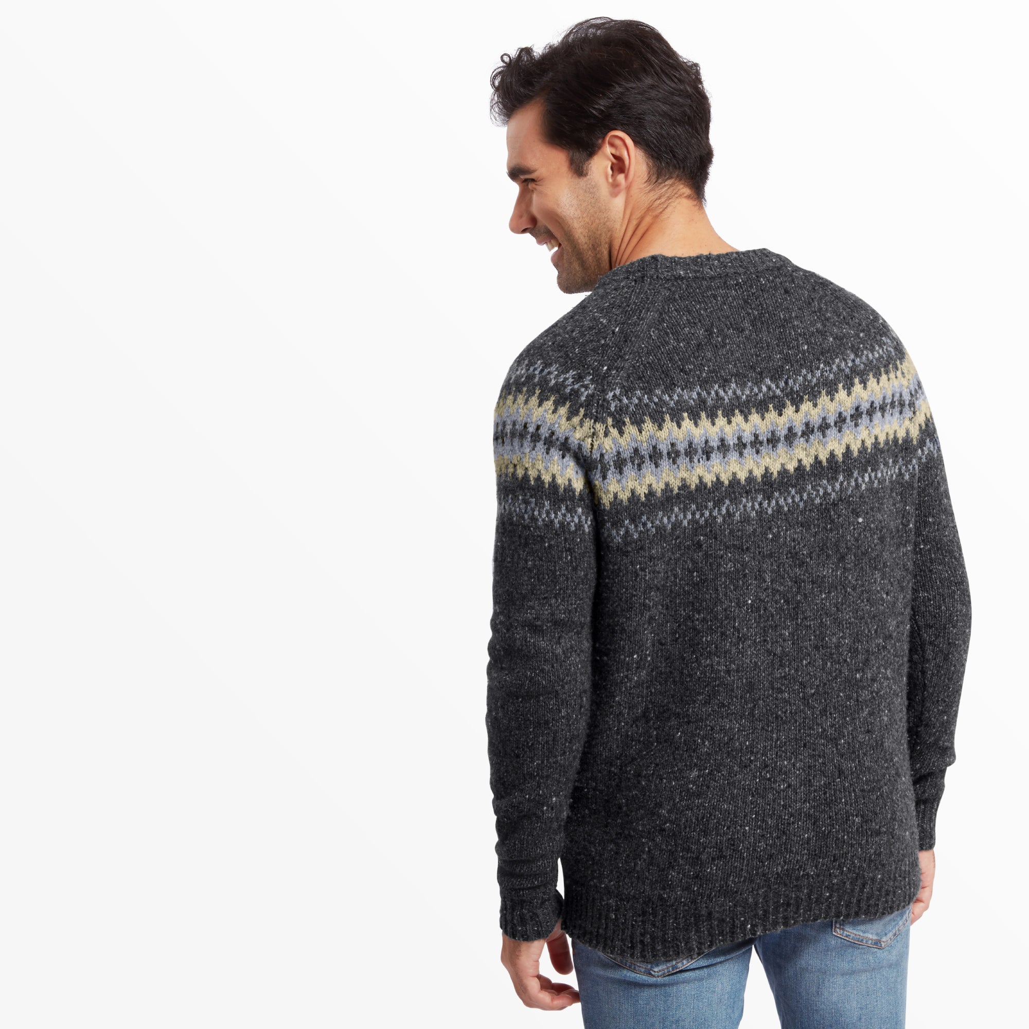 A back view of the male model wearing the Sherpa Adventure Gear Dumji Sweater in Grey, showing the continuation of the Fair Isle pattern across the upper back and sleeves. The ribbed hem and cuffs are visible, and the knit fabric has a subtle textured appearance. The model’s hands are relaxed at his sides, and the light blue jeans and brown boots complete the outfit. The background is plain white.