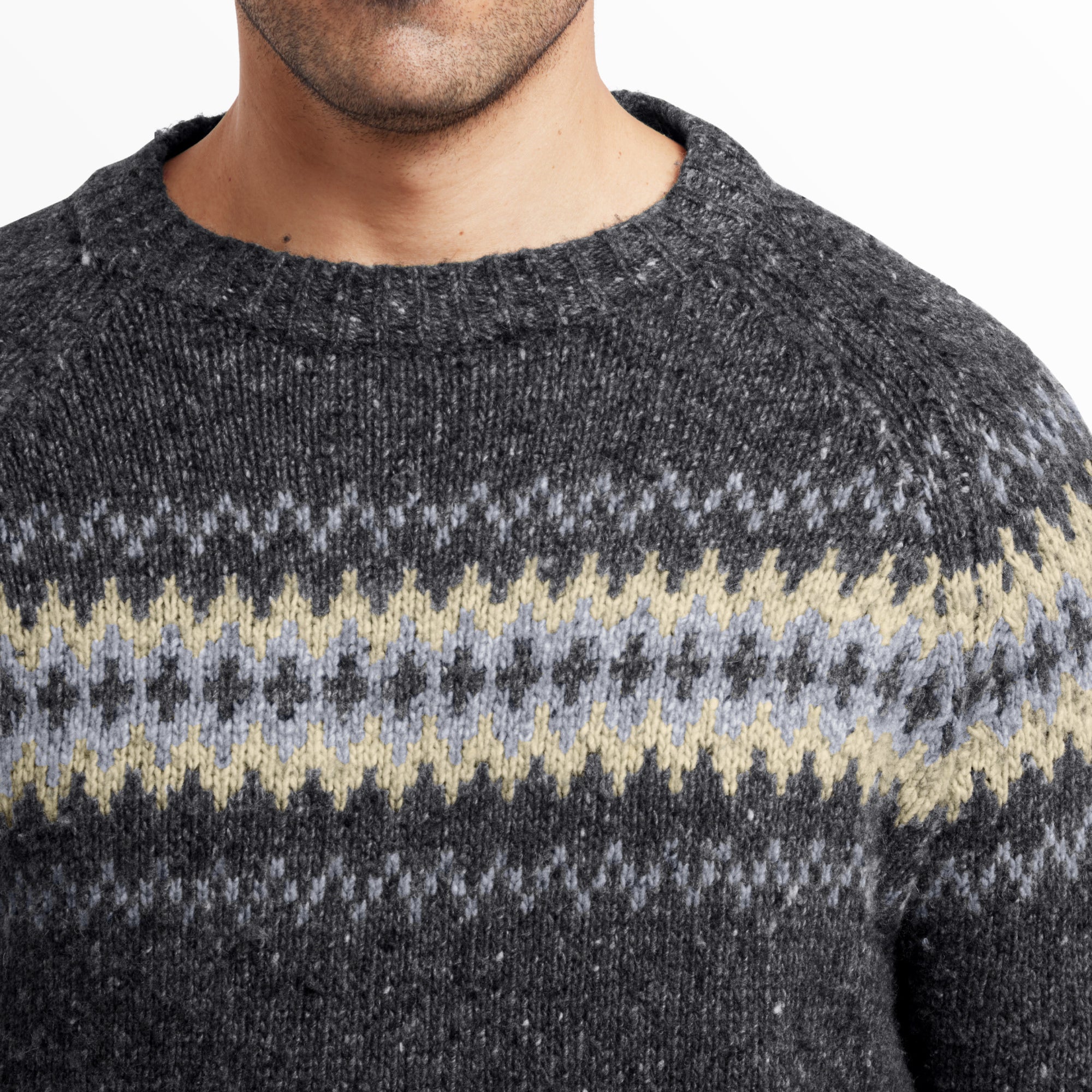 A close-up of the upper chest and shoulders of the Sherpa Adventure Gear Dumji Sweater in Grey, highlighting the intricate knit pattern in light grey, cream, and olive green. The ribbed neckline is also visible, showing the sweater’s texture and knit quality. The background is plain white.