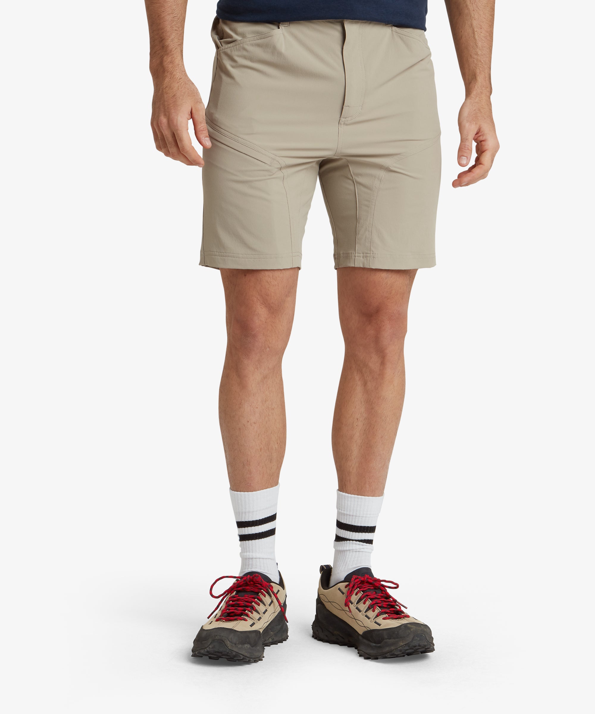 The model is wearing Sherpa Adventure Gear Jamyang Shorts in Beige with a clean front view that highlights the structured waistband, button closure, and front pockets. The model pairs the shorts with white trainers, showcasing a casual style. The background is plain to keep focus on the shorts.