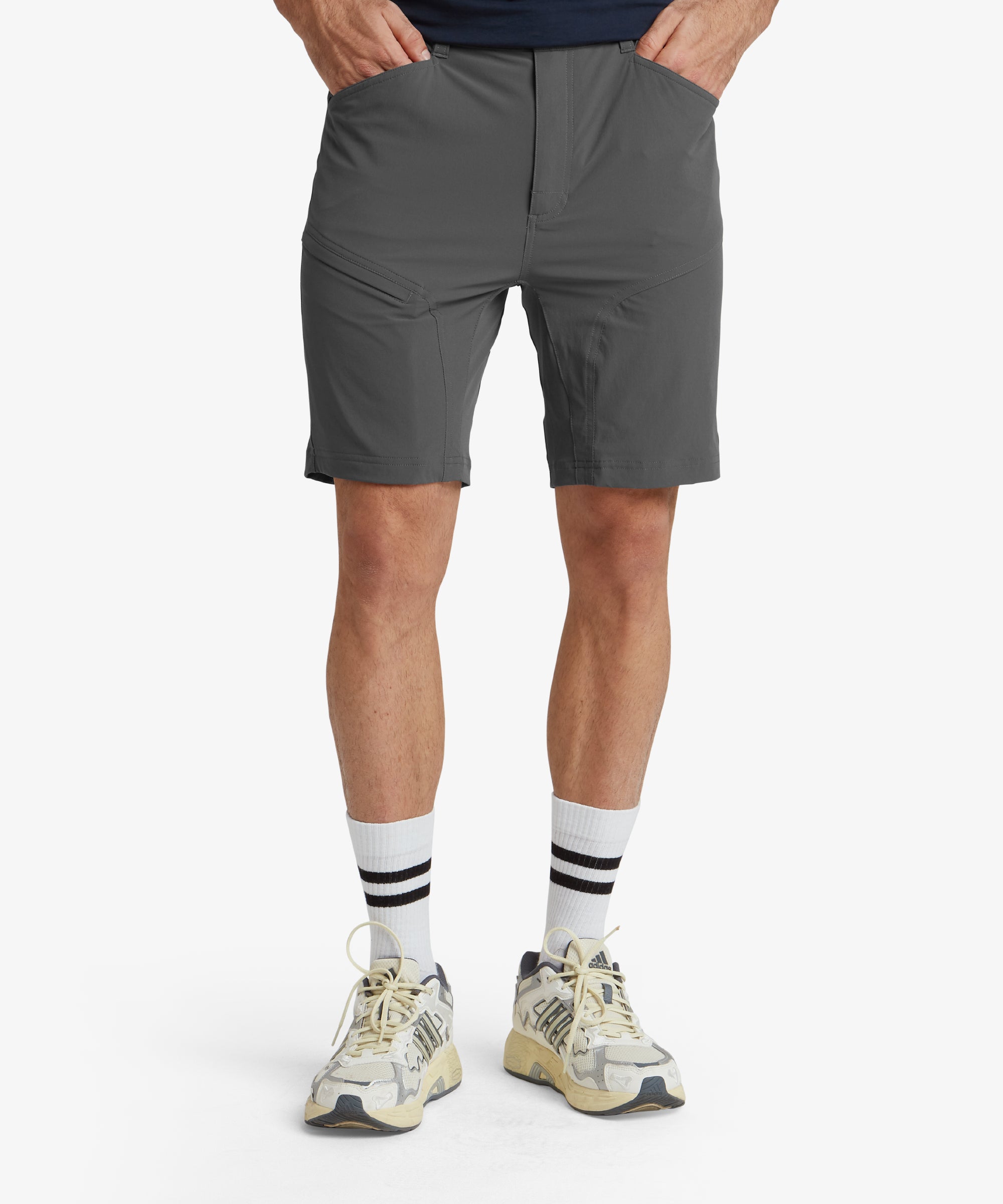 A close-up front view of a pair of Sherpa Adventure Gear Jamyang Shorts in Grey, showcasing the clean design and structure. The shorts are paired with white casual trainers, highlighting their versatile style. The waistband features a button closure and belt loops, suitable for casual or outdoor wear.