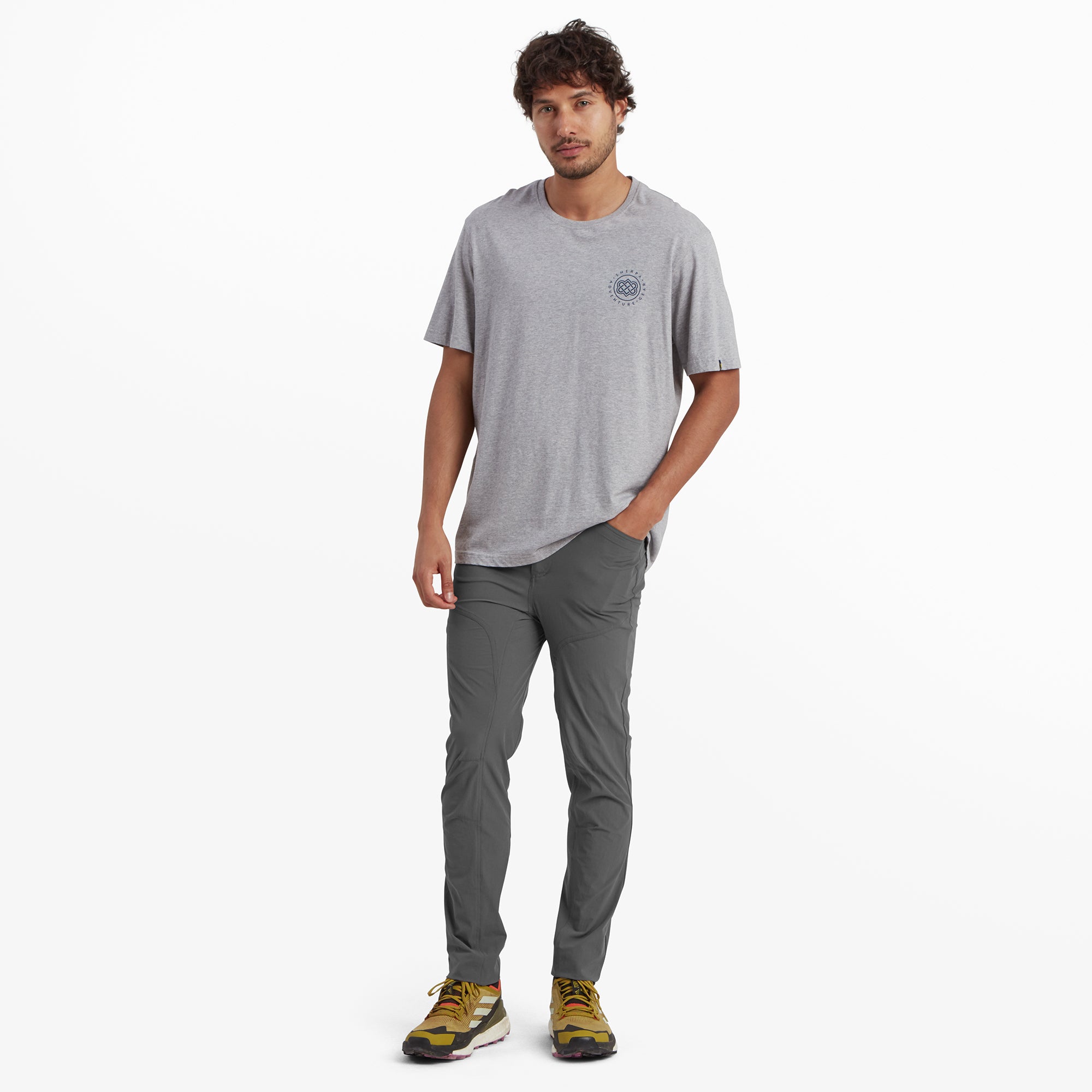 The model is dressed in a complete outfit, wearing the Sherpa Adventure Gear Jamyang trousers in Grey paired with a light gray t-shirt and yellow hiking shoes. This full-length image highlights the overall fit and versatility of the trousers, suitable for outdoor or casual settings.