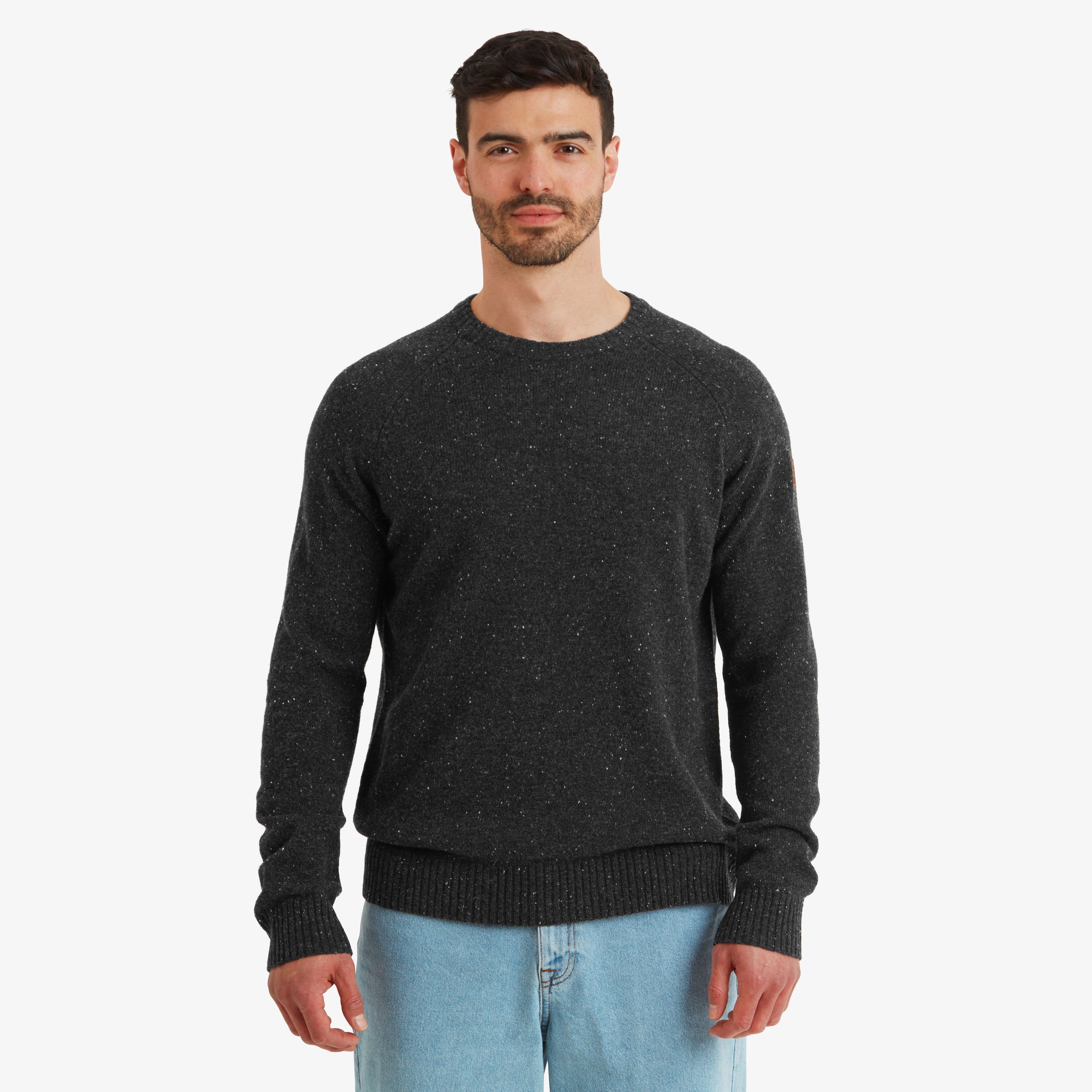 A male model stands confidently wearing a Sherpa Adventure Gear Kangtega Crew Sweater in Black. The sweater features a subtle speckled design for texture. The ribbed hem and cuffs provide a snug fit. The model pairs the sweater with light-wash blue jeans for a casual yet polished look.