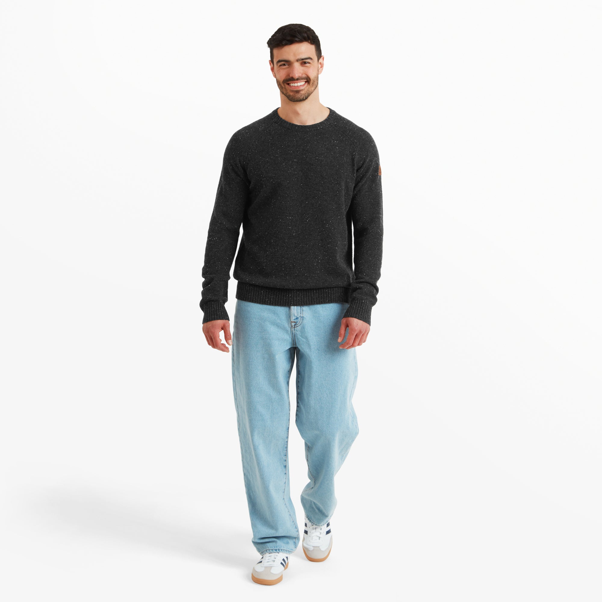 The model is captured mid-step while smiling, showcasing the relaxed and versatile fit of the Sherpa Adventure Gear Kangtega Crew Sweater in Black. The sweater’s lightweight yet cozy material pairs effortlessly with light-wash jeans and white trainers, highlighting its suitability for everyday wear.