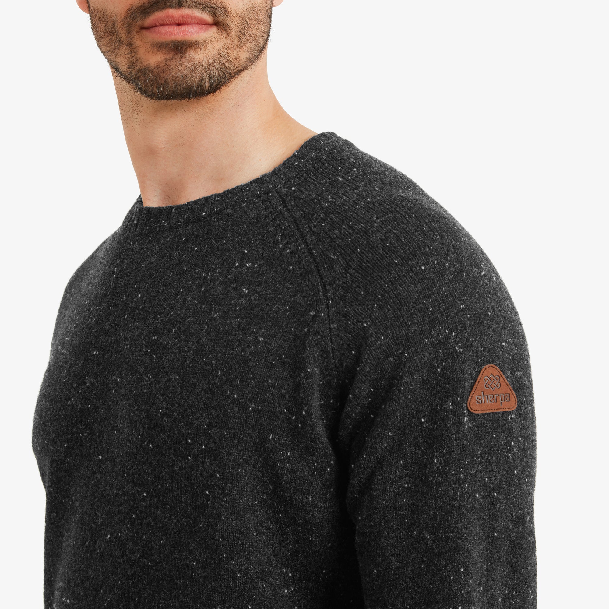 A zoomed-in view of the Sherpa Adventure Gear Kangtega Crew Sweater in Black's left sleeve, highlighting the Sherpa Adventure Gear leather patch logo. The speckled knit fabric is prominently displayed, showcasing the intricate weaving and texture of the material.