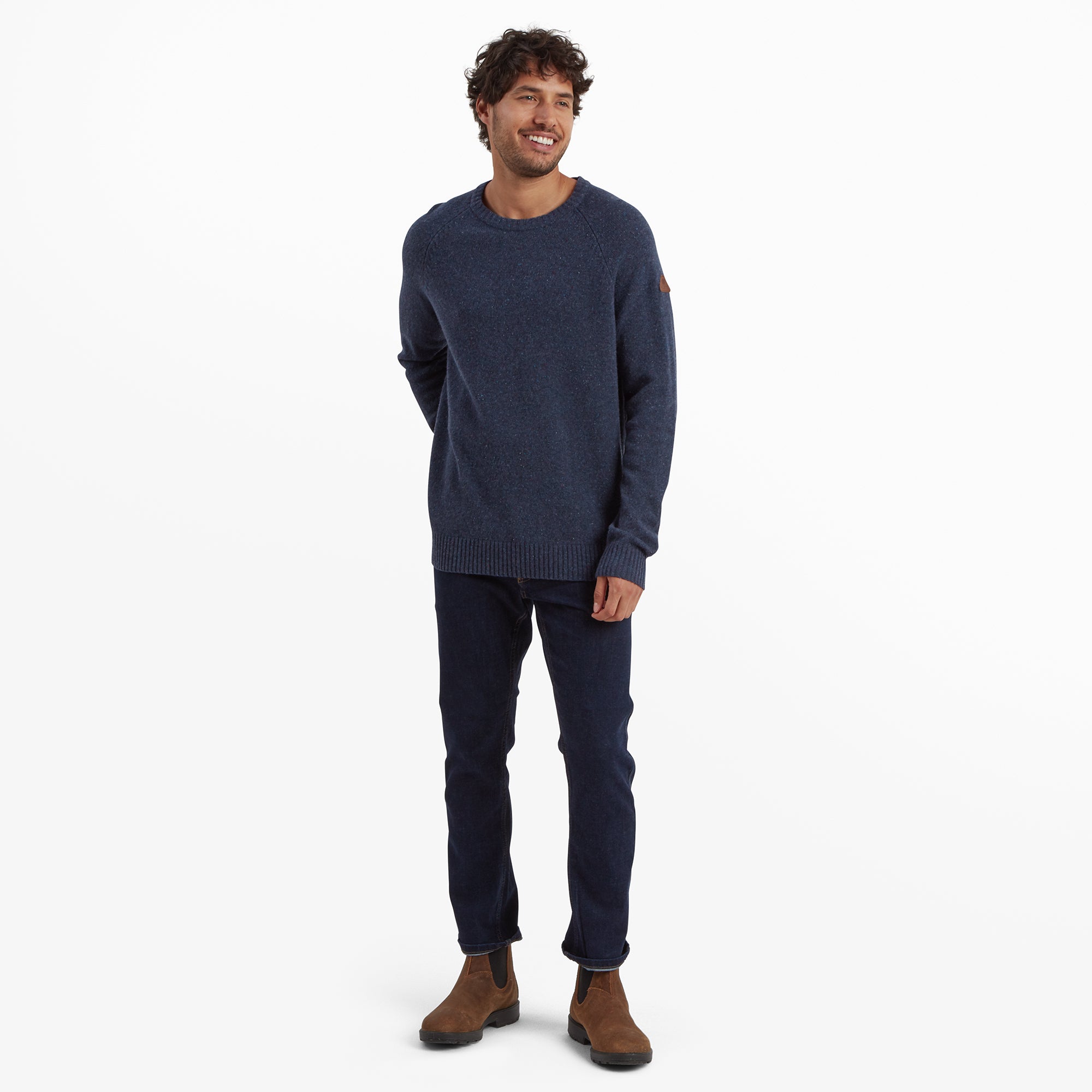 Full-length view of the man modeling the same Sherpa Adventure Gear Kangtega Crew Sweater in Blue, styled with dark denim jeans and brown Chelsea boots. He is smiling, exuding a relaxed and approachable vibe.