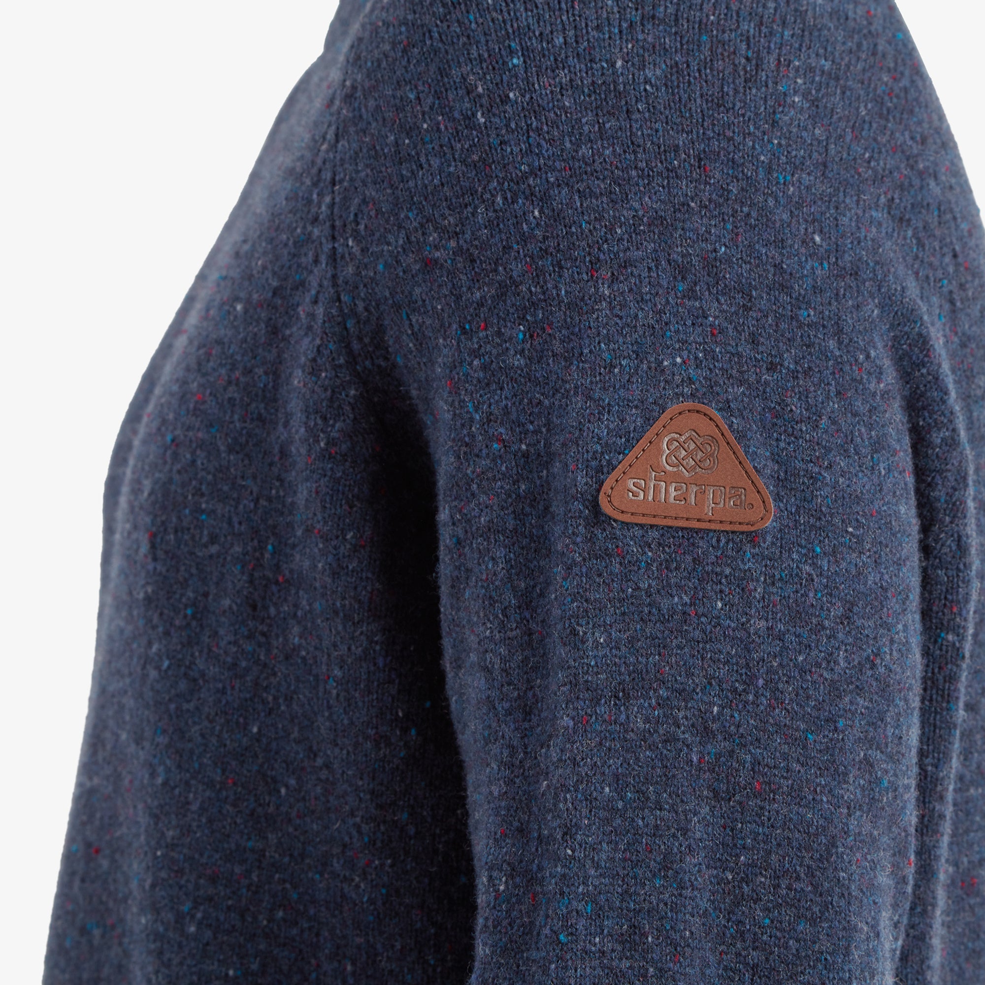 Close-up of the left sleeve, displaying the textured Sherpa Adventure Gear Kangtega Crew Sweater in Blue in detail and a leather triangular patch embossed with the Sherpa logo for a touch of craftsmanship and branding.